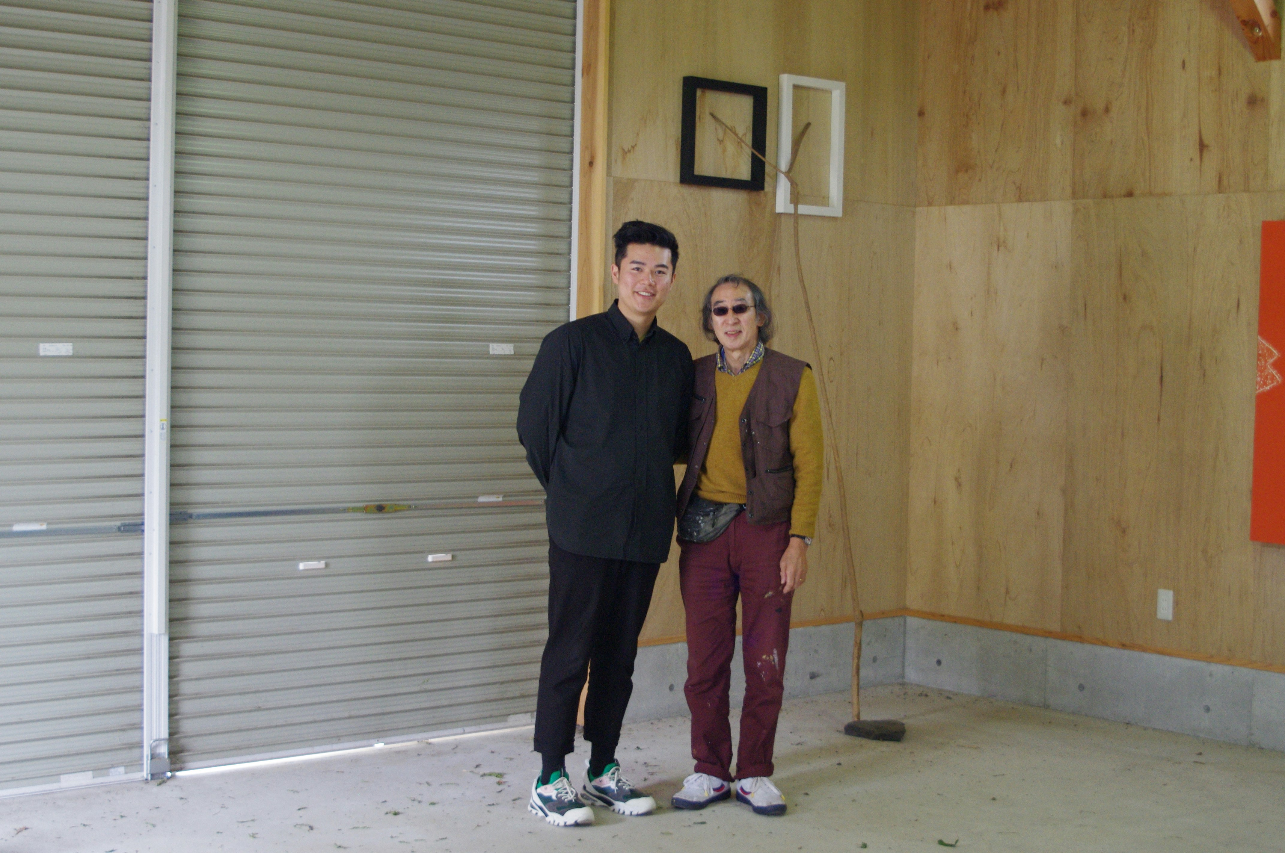 A Chinese-Australian man in all-black and white sneakers, standing next to Kishio in his studio, both smiling at the camera.