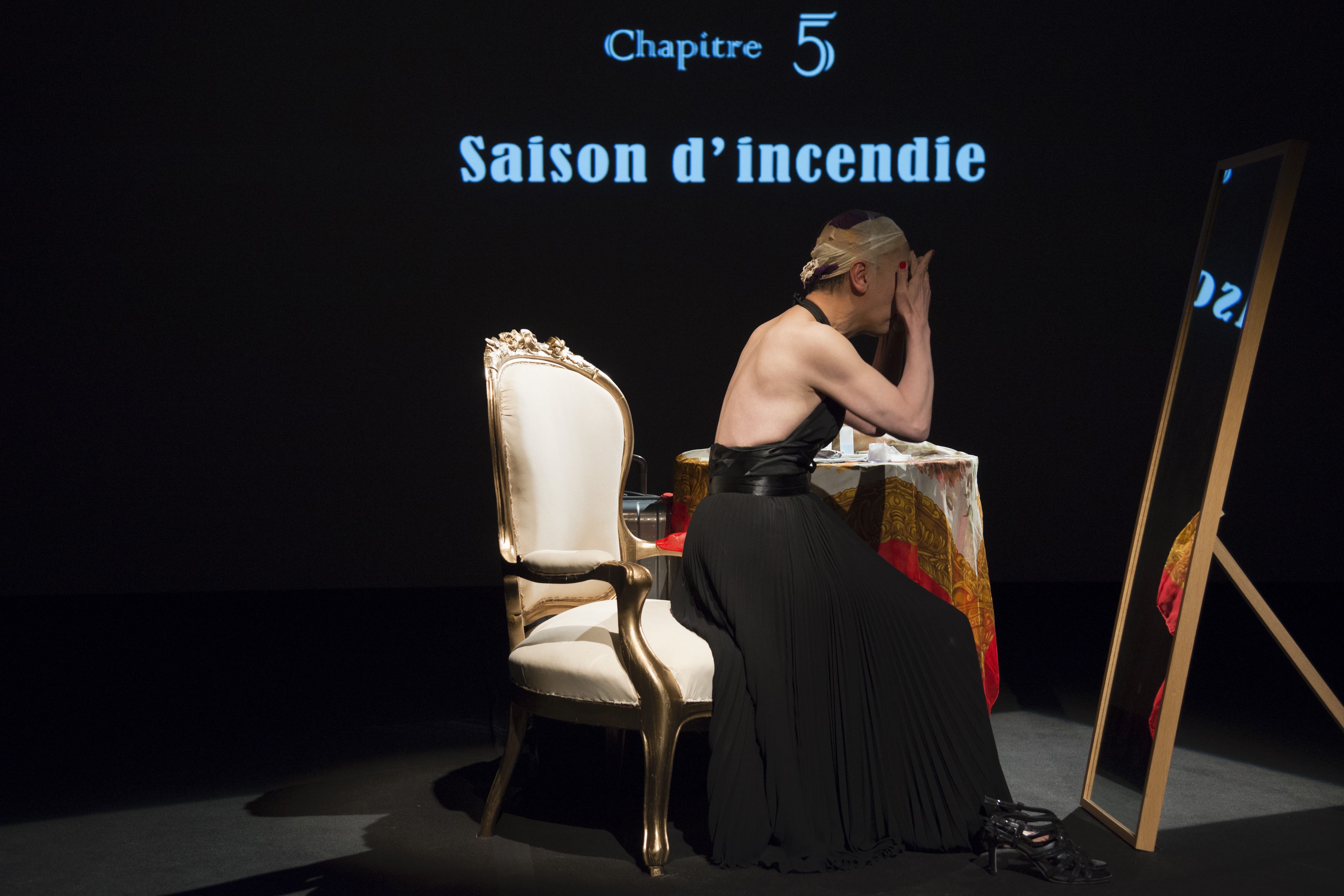 A muscular figure in a black floor-length halter dress sitting in an ornate armchair with their hands over their face and their head taped in a beige fabric. They are facing a floor-length mirror, with the words "Chapitre 5: Saison d'incendie" projected in blue behind them.