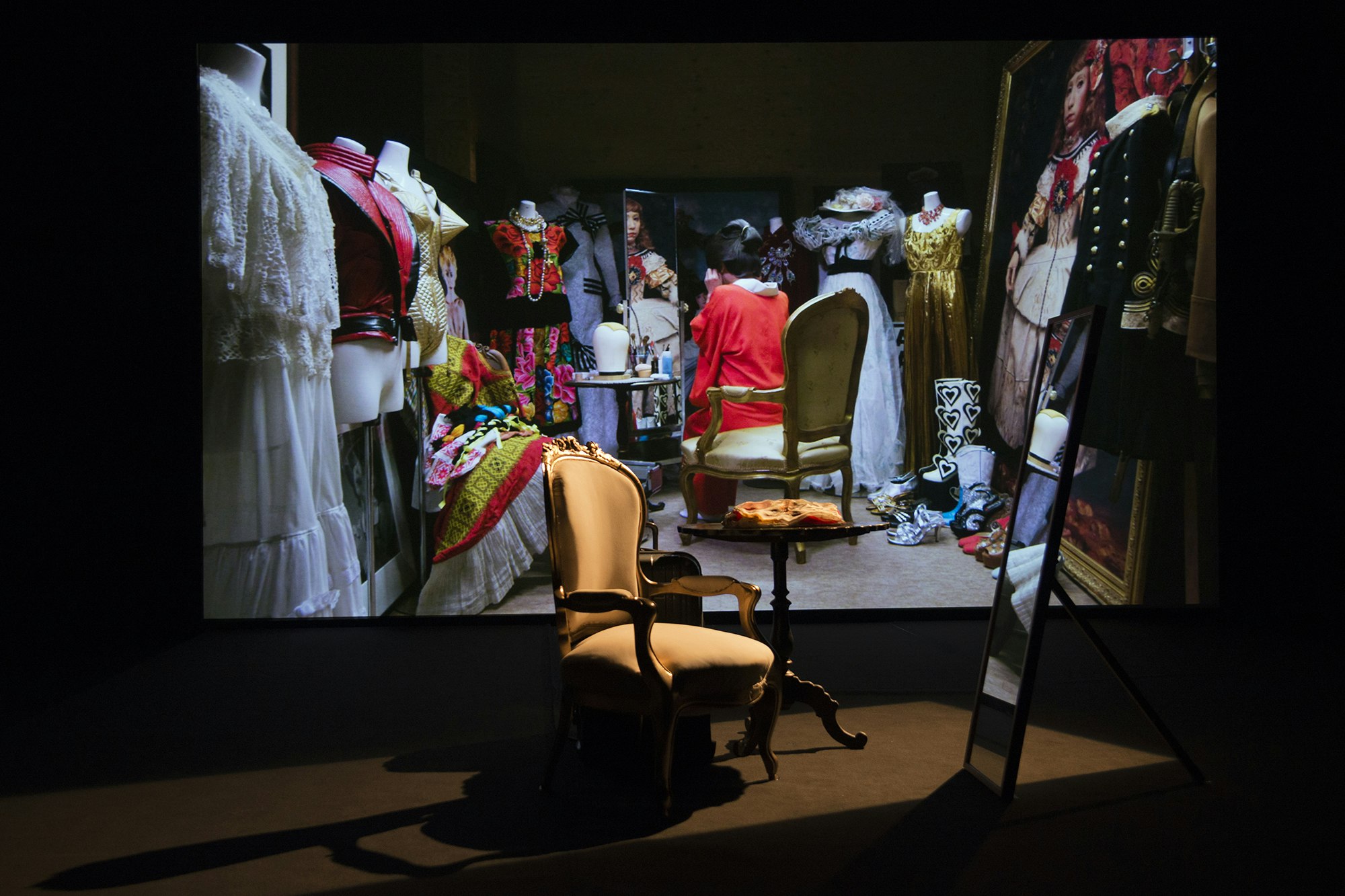 A yellow armchair under a spotlight, next to a wooden table and mirror. Behind is a projection of a figure dressed as a Japanese geisha with their back to us, surrounded by mannequins dressed in floral dresses, golden suits and floor-length dresses, with a Yasumasa Morimura painting of a girl in historical dress framed in gold in the corner.