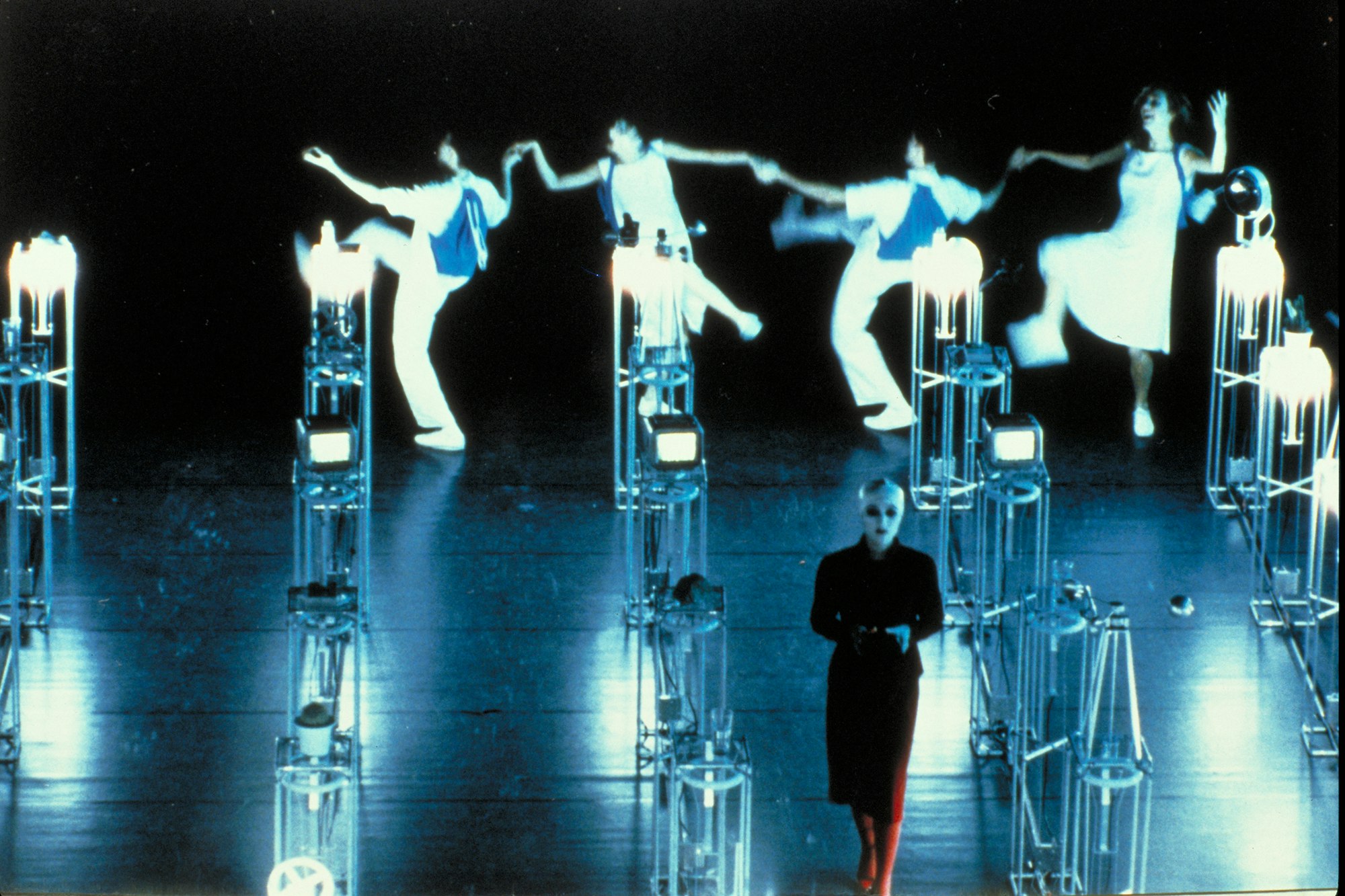 A negative still of four individuals holding hands and dancing joyfully, while a figure clothed in a long black dress walks away from the group.