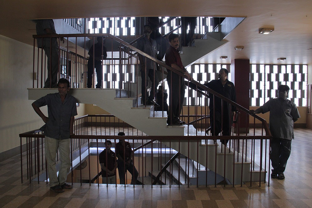 A multi-level staircase, where multiple brown-skinned male-presenting figures in grey button-ups and dark pants stand on different parts of the staircase. 