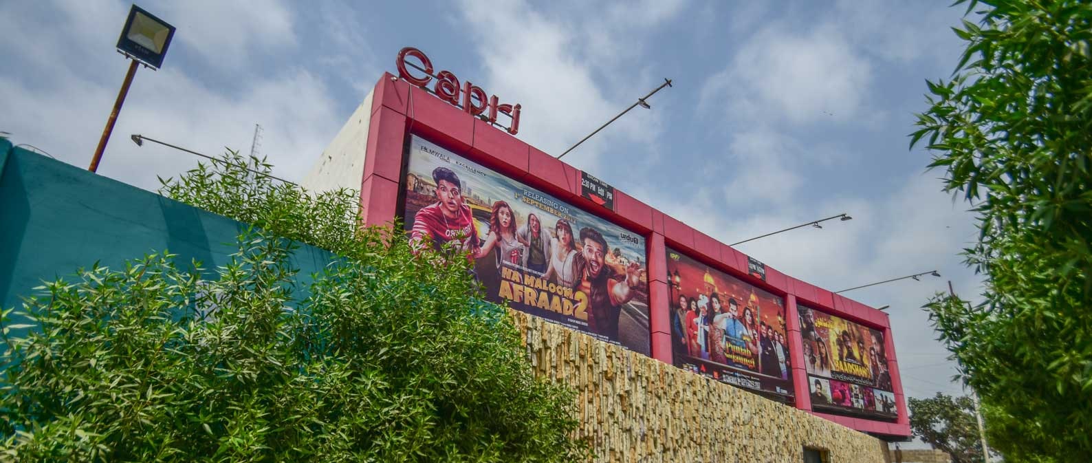 The pink facade of a cinema posted with three large colourful movie posters, surrounded by greenery.