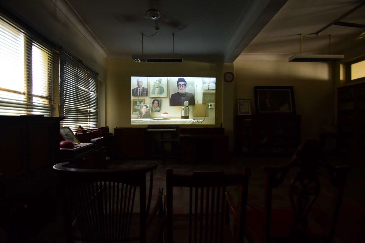 Chairs facing a projector screen in a dark room with closed blinds. The screen shows various photos of older male figures, some wearing glasses, some in traditional dress.