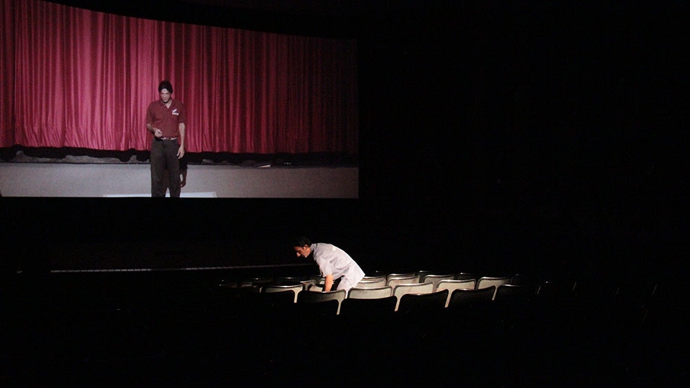 A male-presenting figure leaning down in a row of cinema seats while the cinema screen shows a figure in a red dress shirt and black trousers standing in front of a floor length red curtain.