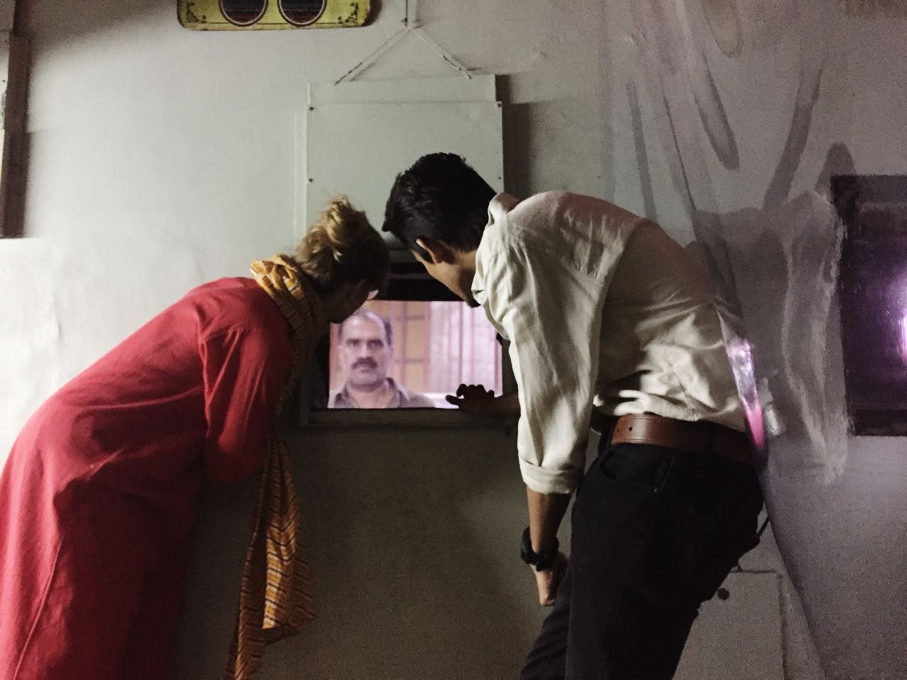 A female-presenting figure in a yellow striped scarf and long red dress, and a male-presenting figure in a white button-up shirt and black trousers, both leaning over to watch a small video screen of a man's face.