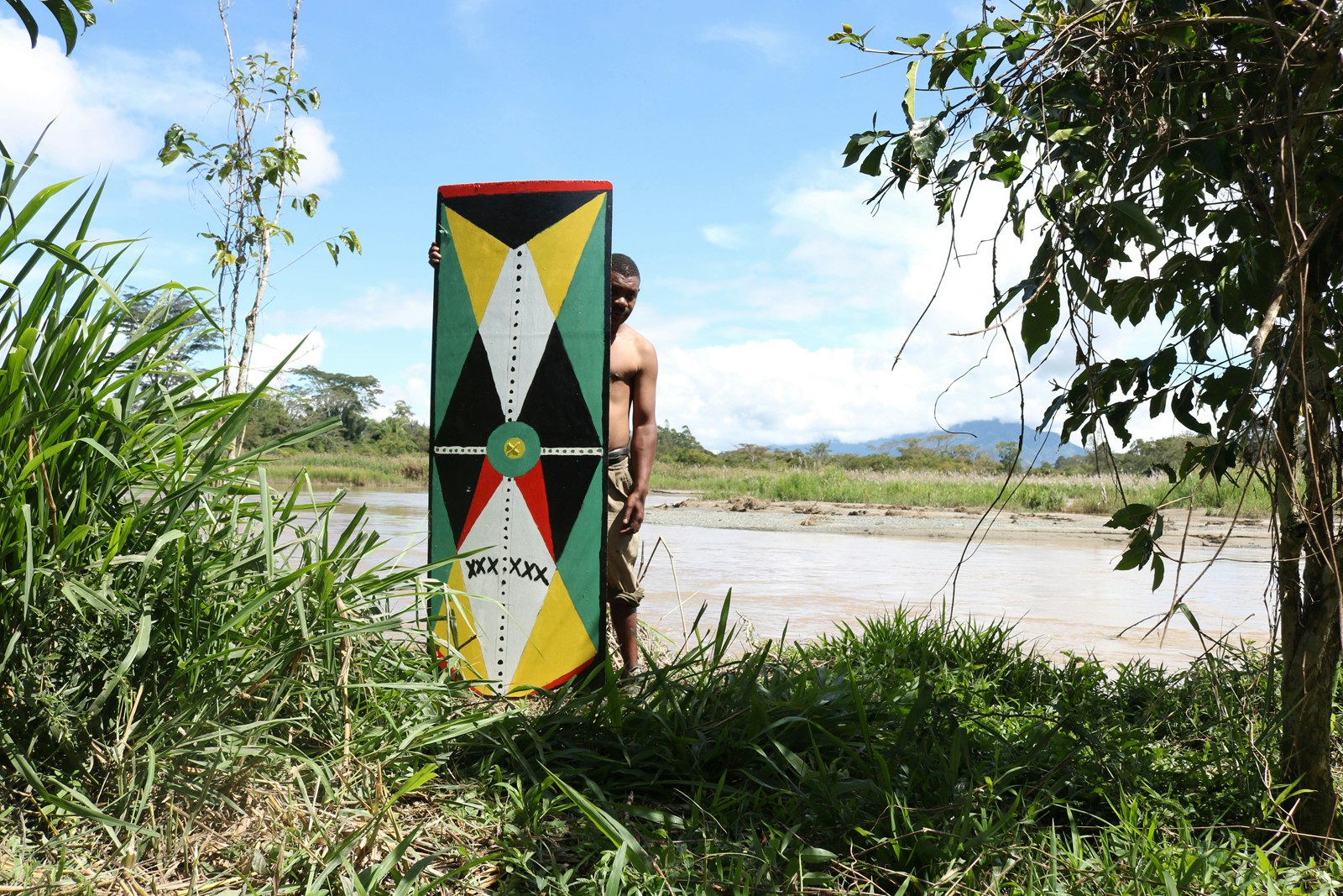 A Pasifika man stands behind a kuman (shield) painted with a geometric pattern of green, yellow, white, red and black shapes. He is standing amongst vegetation on a river bank.