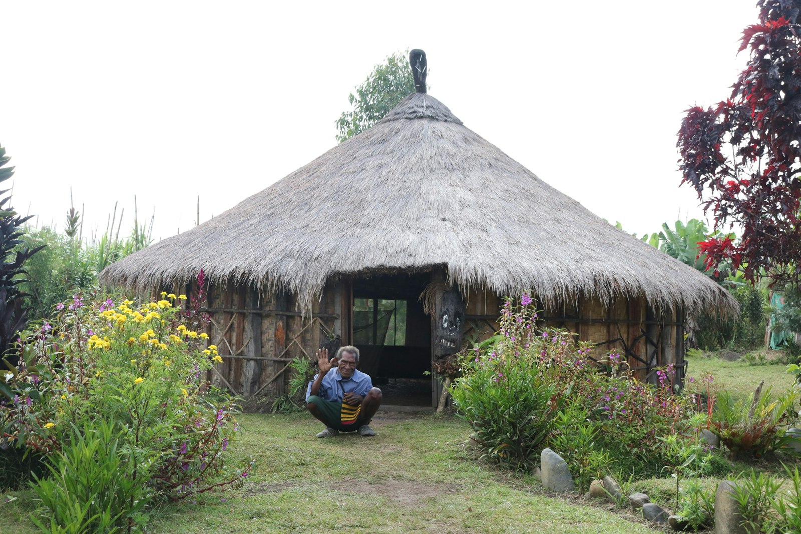 An elderly Pasifika man with dark skin squats outside a grass hut and waves at us, positioned on a pathway between rows of planted pink and yellow flowers.