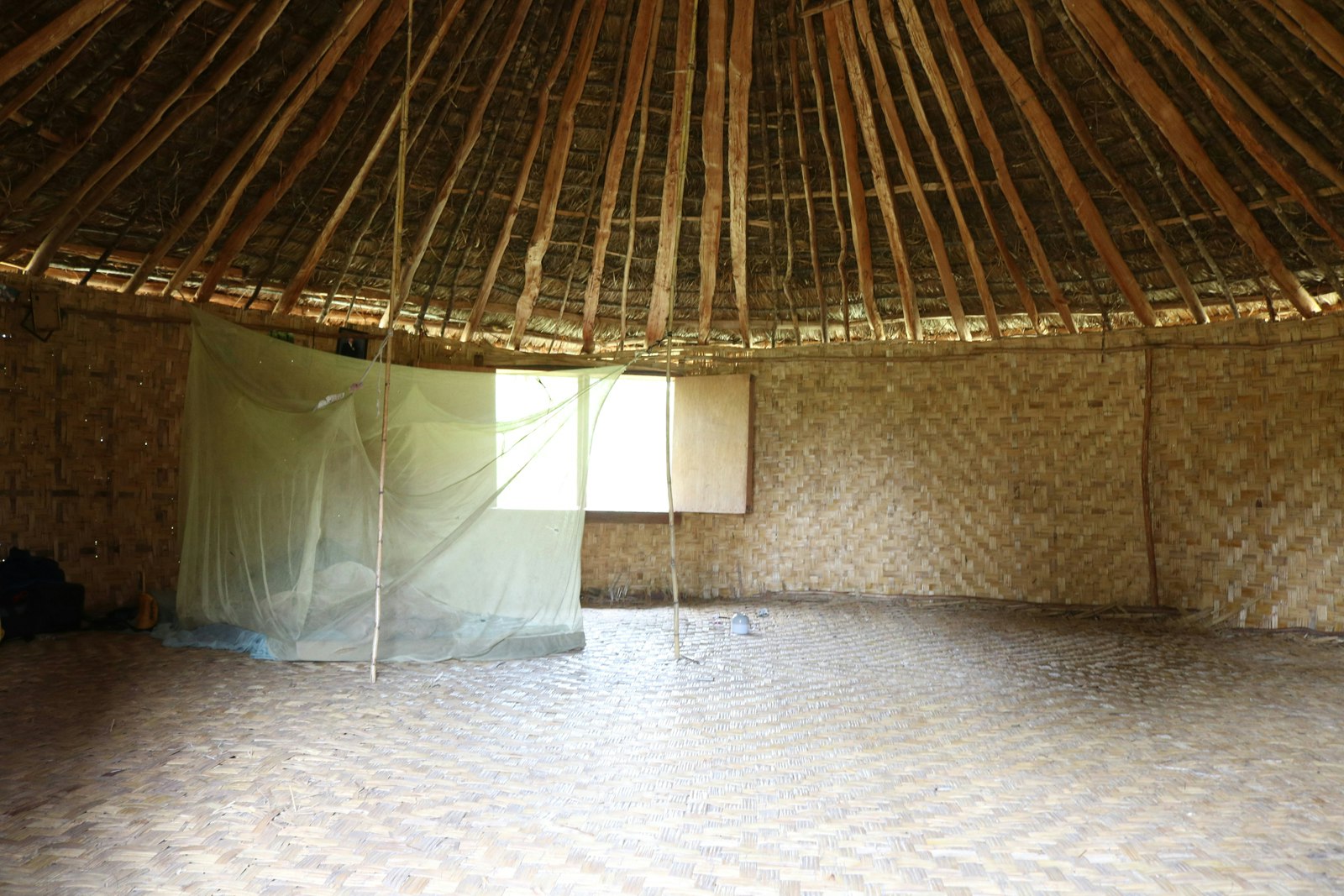 Inside the central room of a grass hut with a pointed roof. The walls and floor are woven from fibre, and there is a window cut into one of the walls.