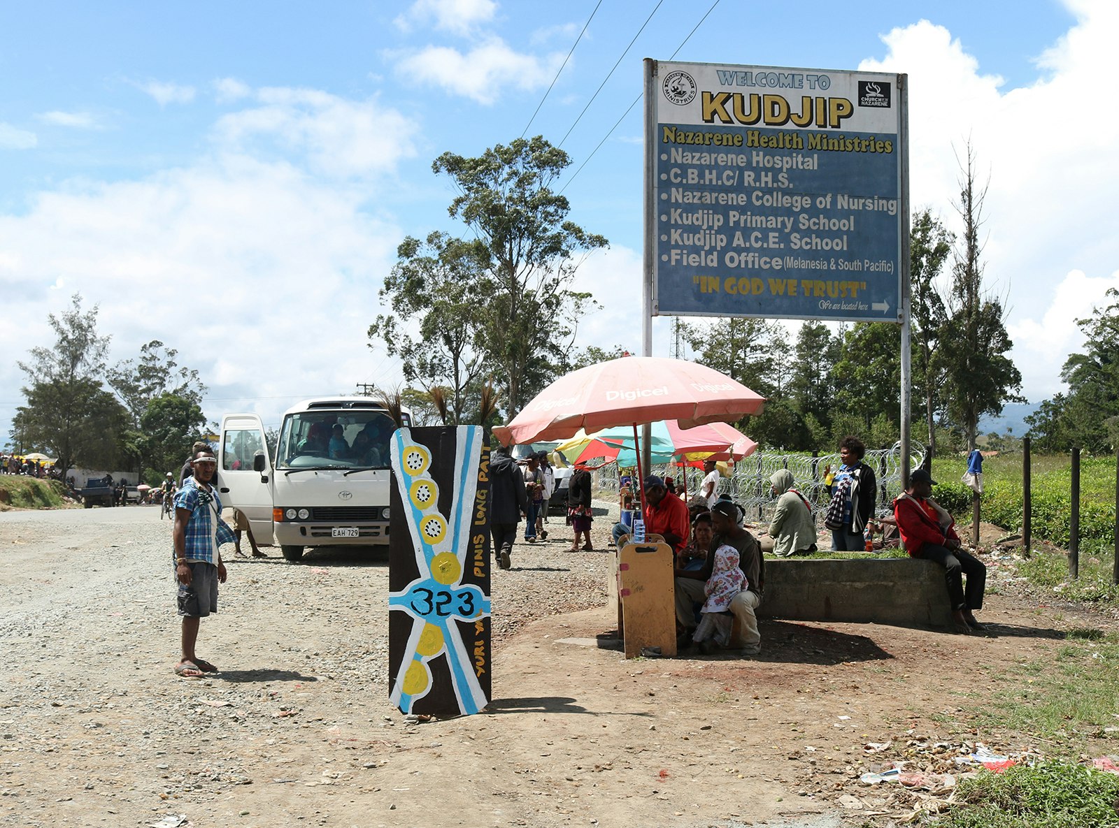 A dirt road in Papua New Guinea with a blue and yellow kuman (shield) planted in front of some roadside vendors set up under umbrellas. A sign above them reads, "Welcome to Kudjip... In God We Trust".