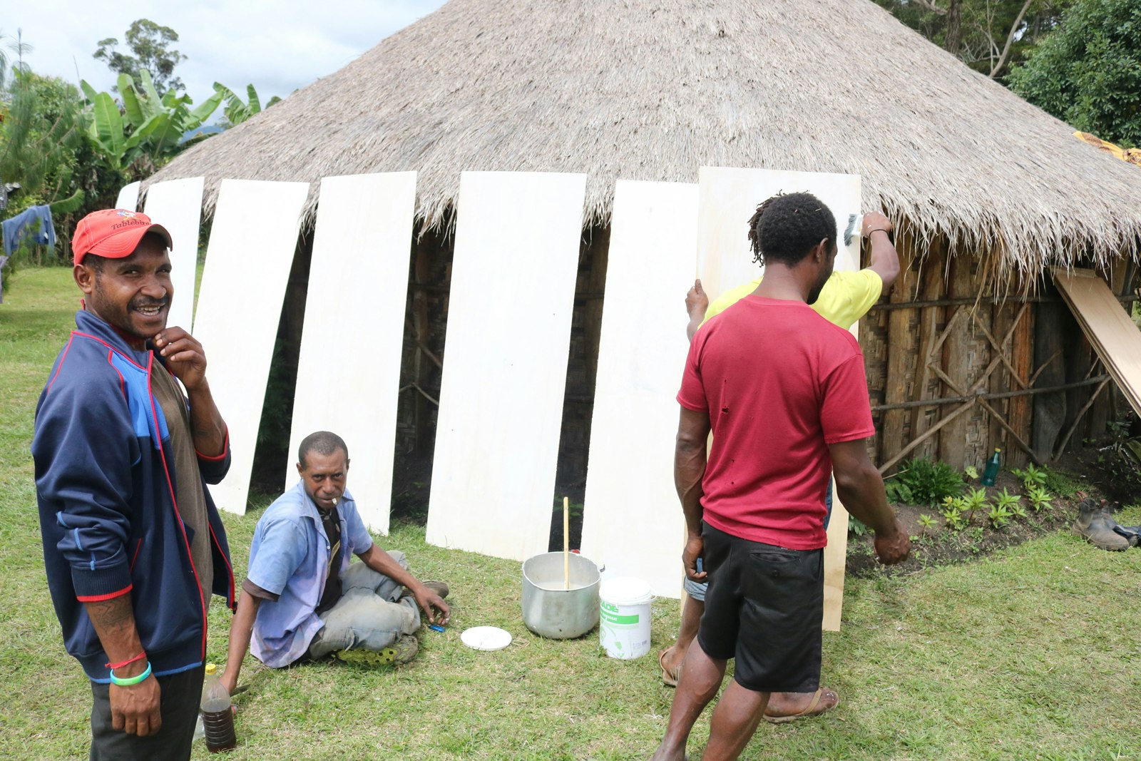 Four Pasifika men working on white plywood sheets stacked against the exterior of a grass hut. The man on the left is smiling with his hand at his chin, mid-speech.