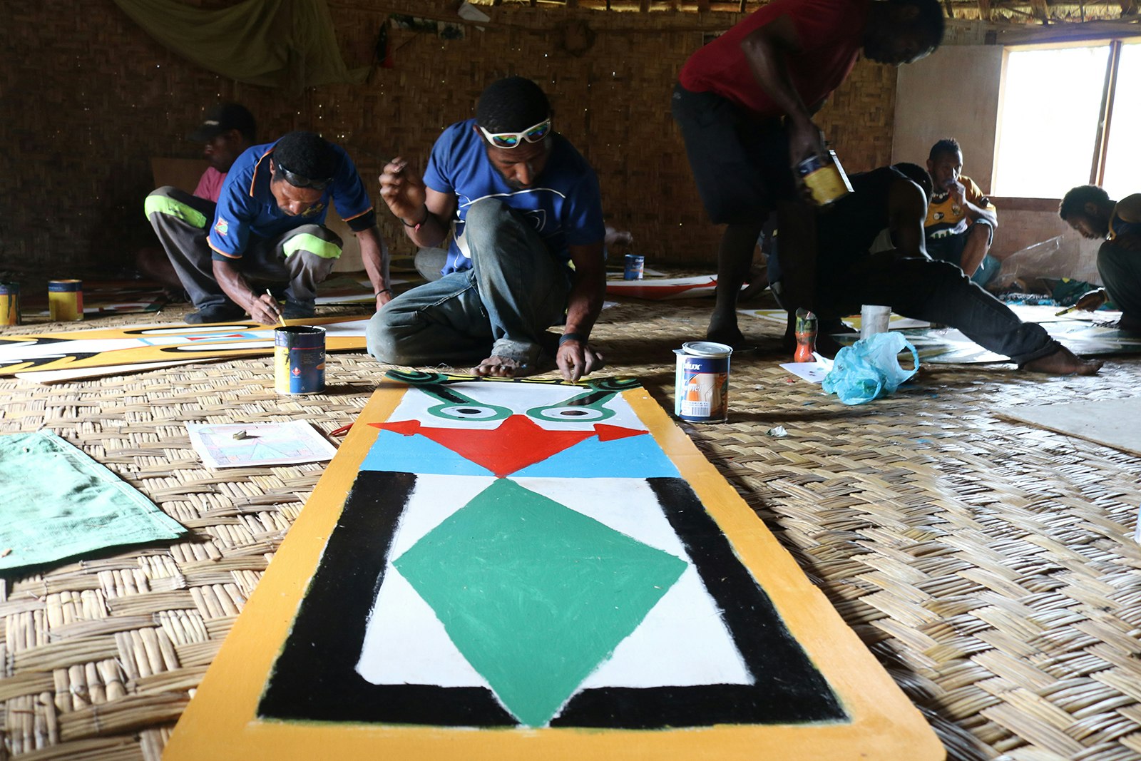 A group of dark-skinned Pasifika men are crouched over sheets on plywood on the floor of a grass hut, painting colourful shapes on each one.