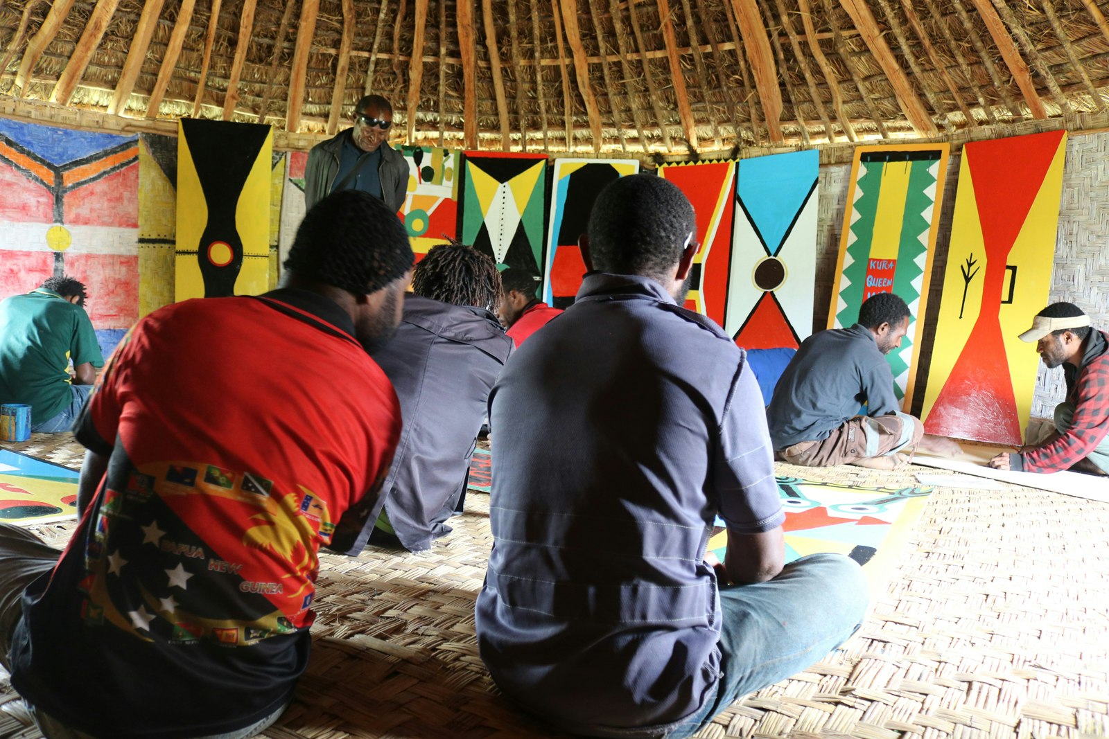 A group of dark-skinned Pasifika men sit cross-legged on the floor of a grass hut, facing a row of kuman (shields) painted in bright colours and leaning against the interior wall of the hut.