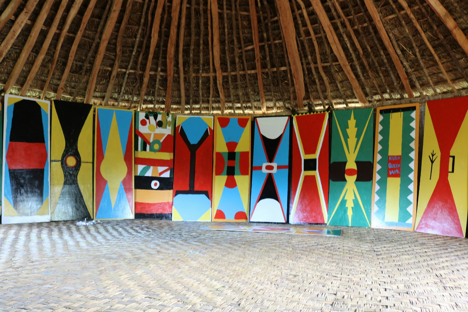 A row of kuman (shields) painted in different geometric shapes and bright reds, yellows, blues and greens. They are lined up in a row inside a grass hut, stretching from the floor to the base of the roof.