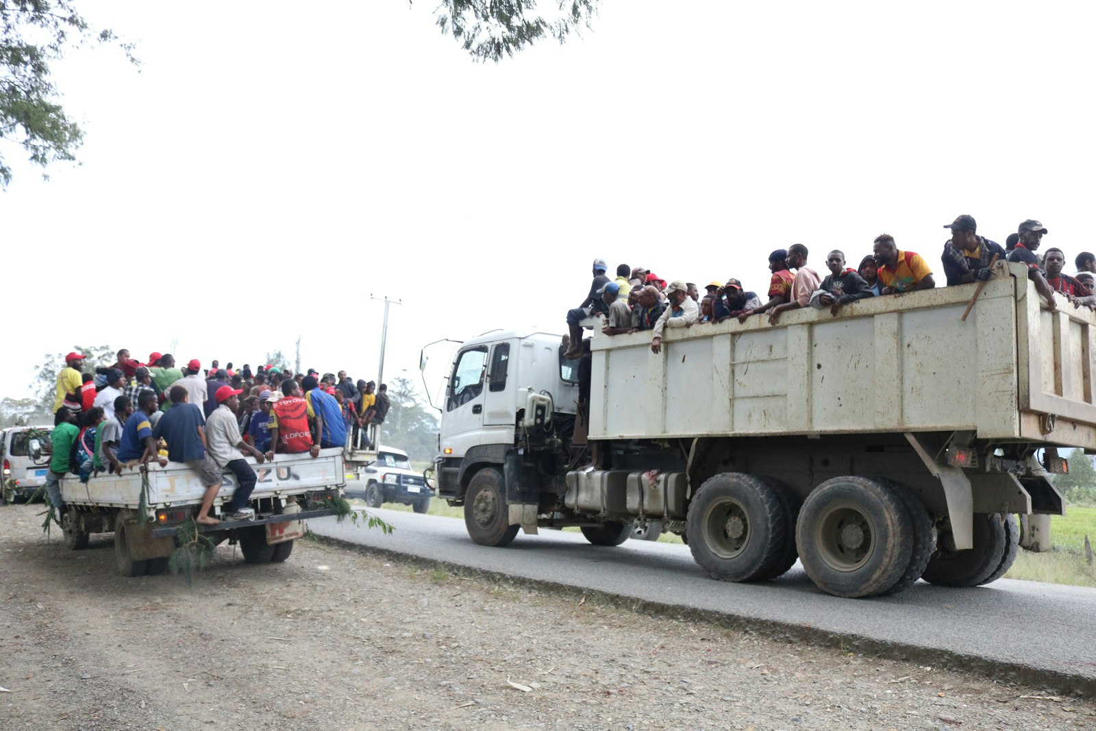 A pickup truck and a dump truck with crowds of Pasifika men piled on the back of each, driving down a narrow dirt highway in Papua New Guinea.