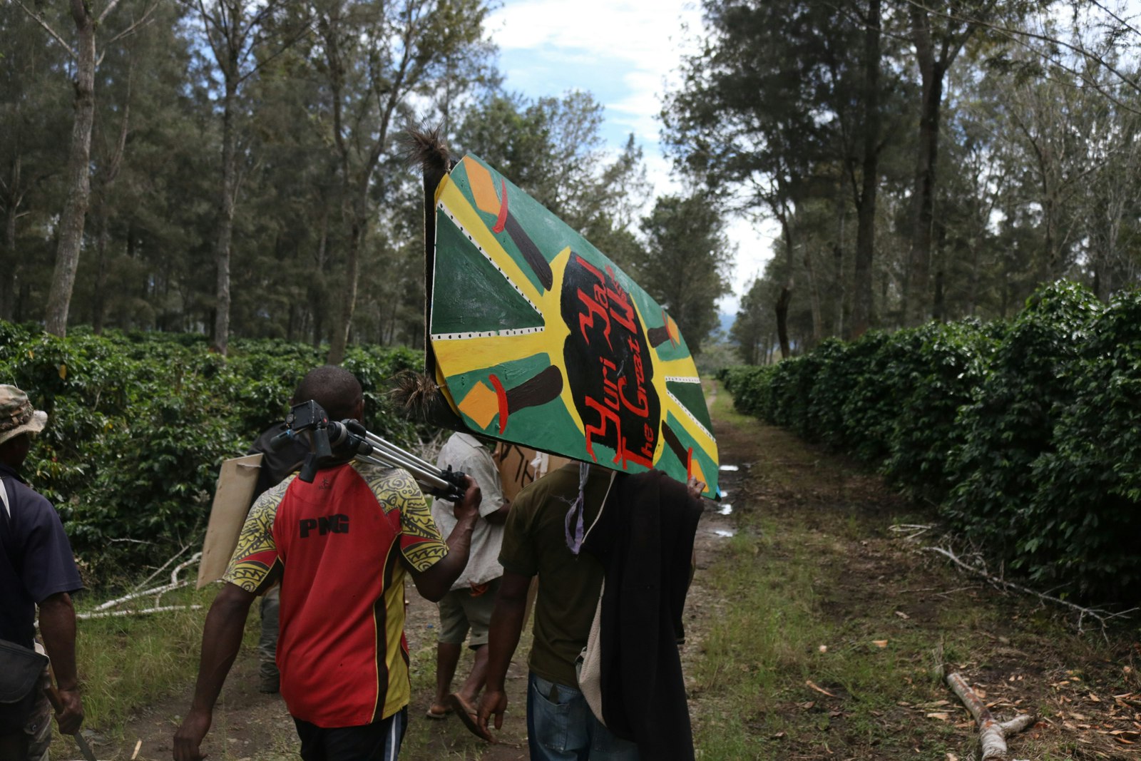 Dark-skinned Pasifika men carrying photography equipment and a green kuman (shield) over their shoulders while walking down a forested path.