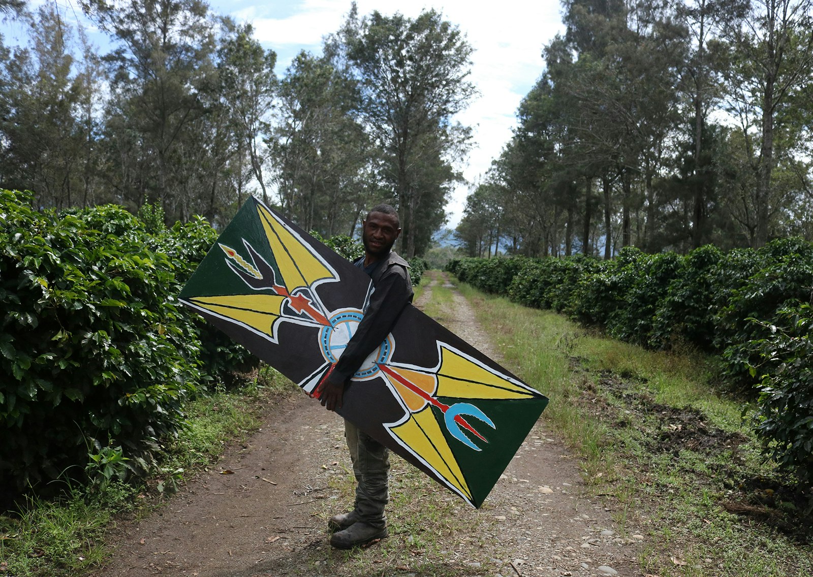 A dark-skinned Pasifika man smiles at the camera while he holds a kuman (shield) painted with yellow arrows and other shapes.