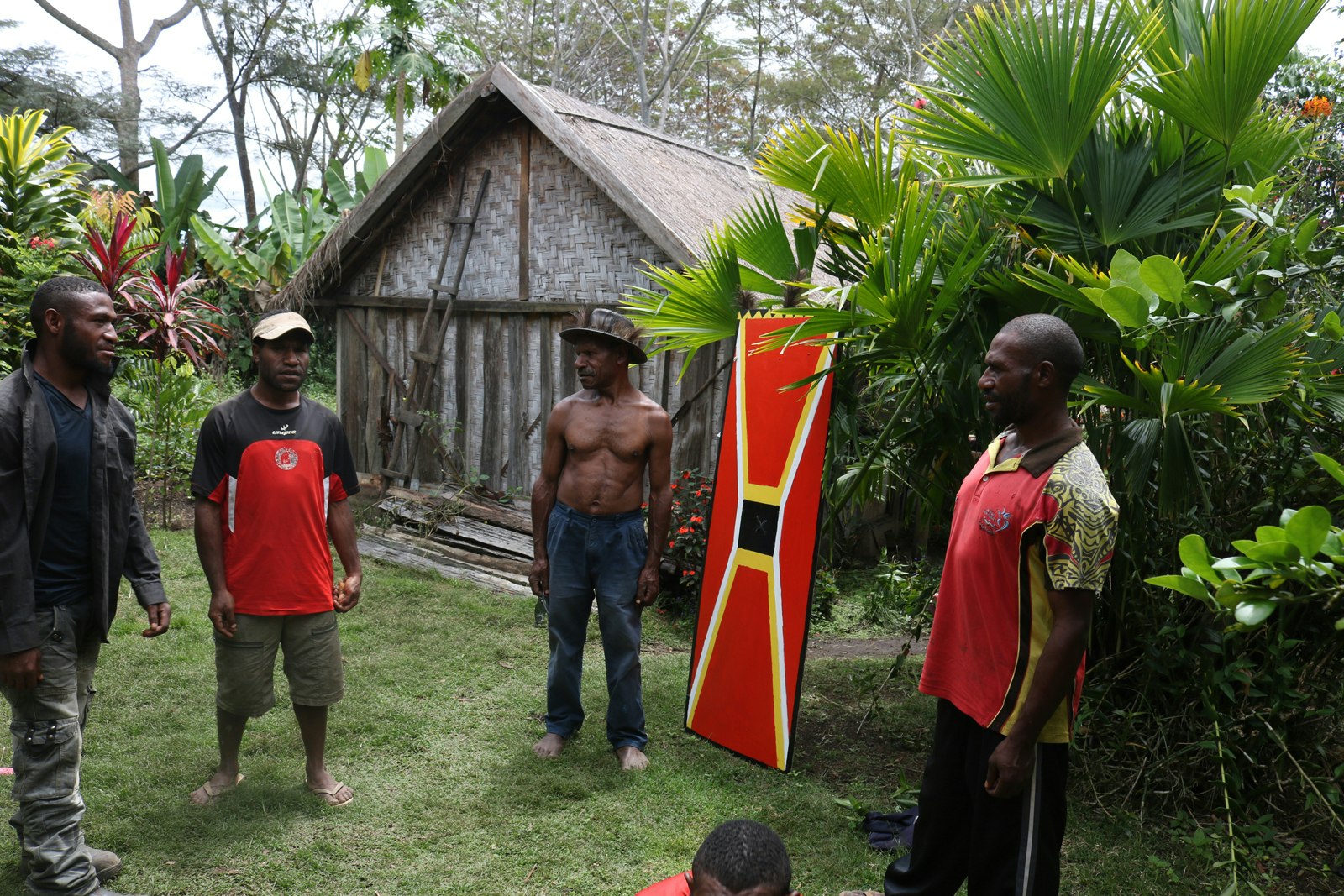 Four dark-skinned Pasifika men, some dressed in bright red shirts, one wearing a jacket and the other shirtless and wearing a hat, stand outside a thatch house, surrounding a red kuman (shield) painted with white and yellow lines. 