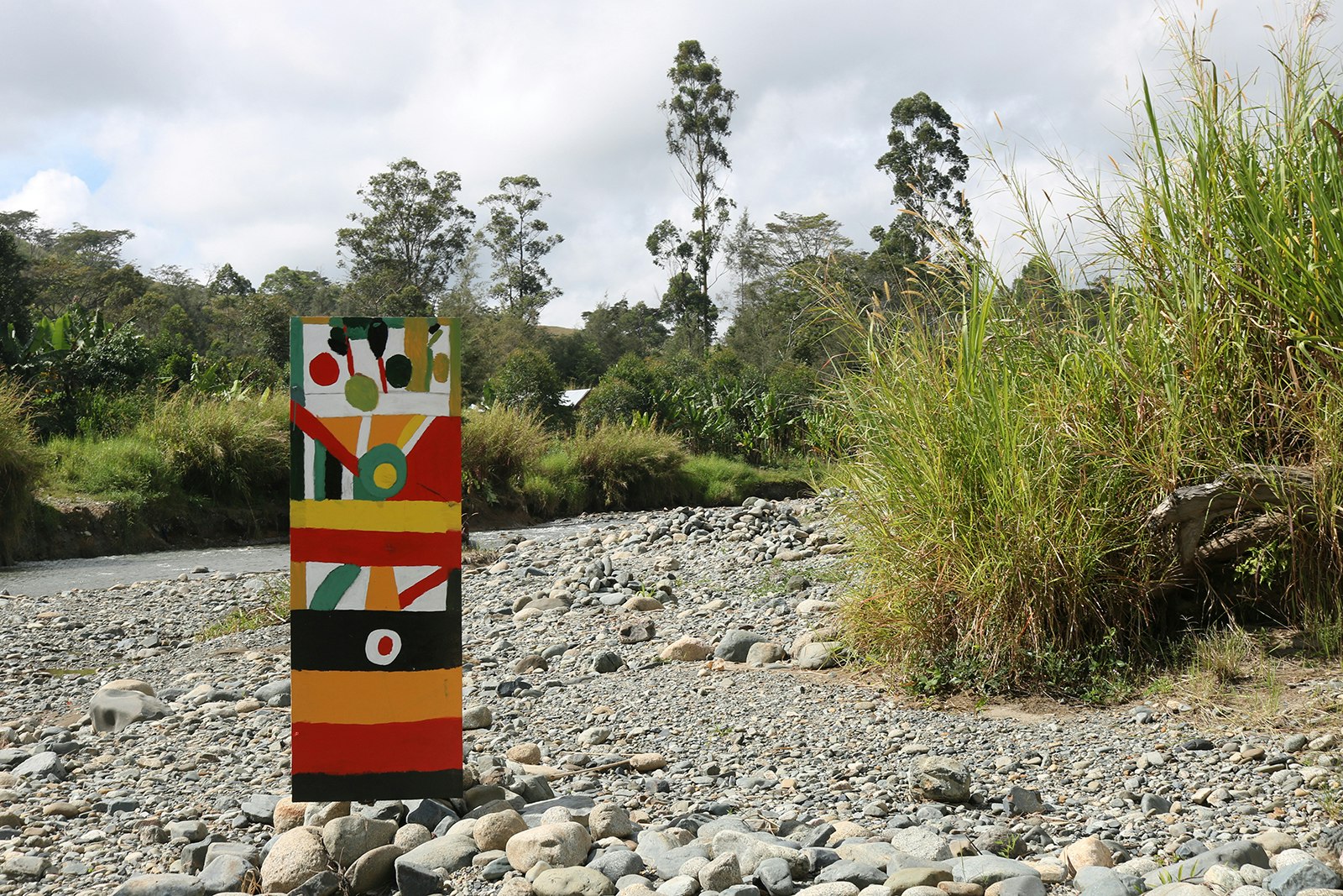 A kuman (shield) painted with a pattern of red, orange, green, yellow, white and black, standing on rocky ground near some tall grass.