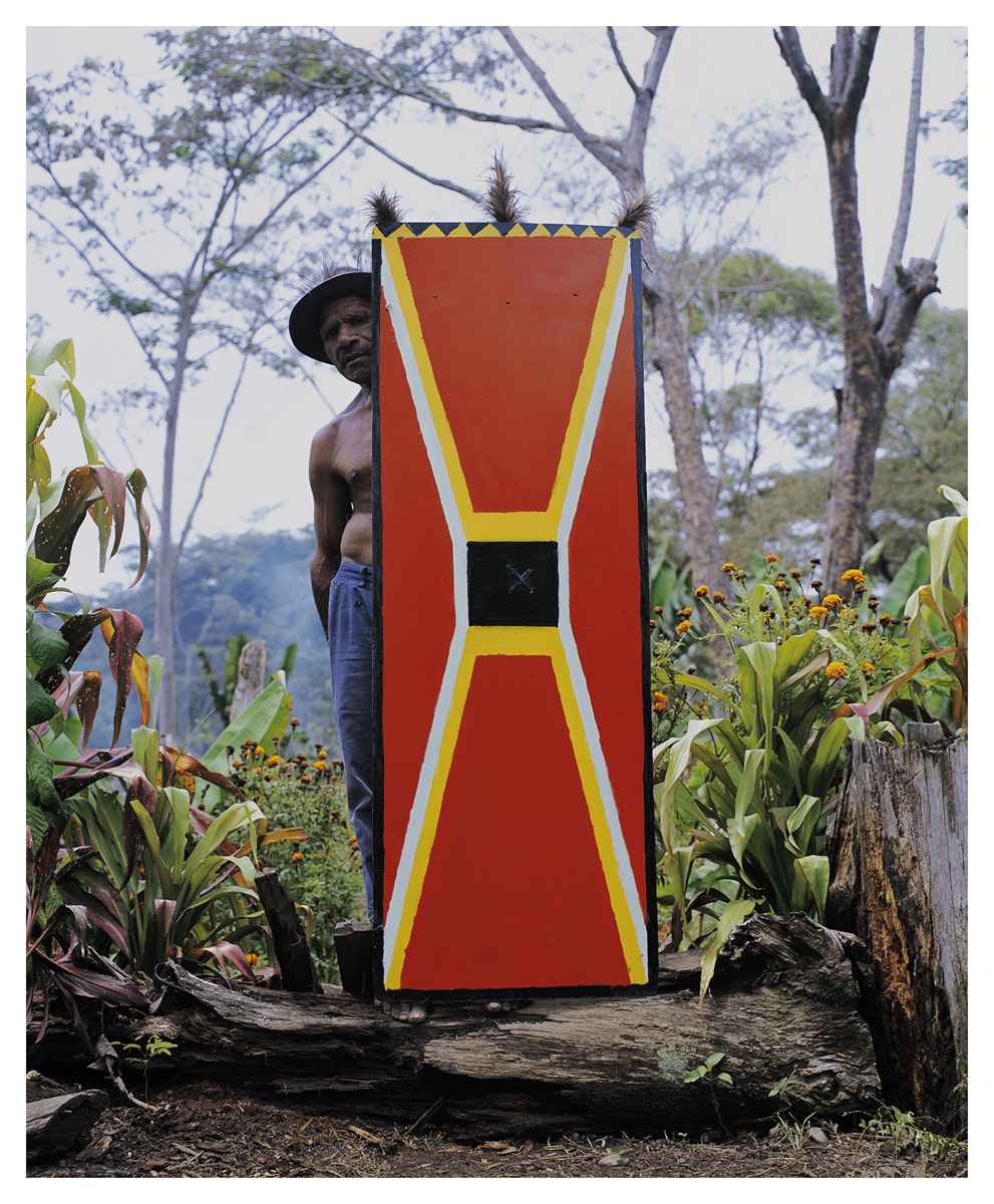 A shirtless Pasifika man wearing trousers and a hat stands behind a bright red kuman (shield) painted with white and yellow diagonal lines. He rests the kuman on a log, standing amongst bushes of orange flowers.