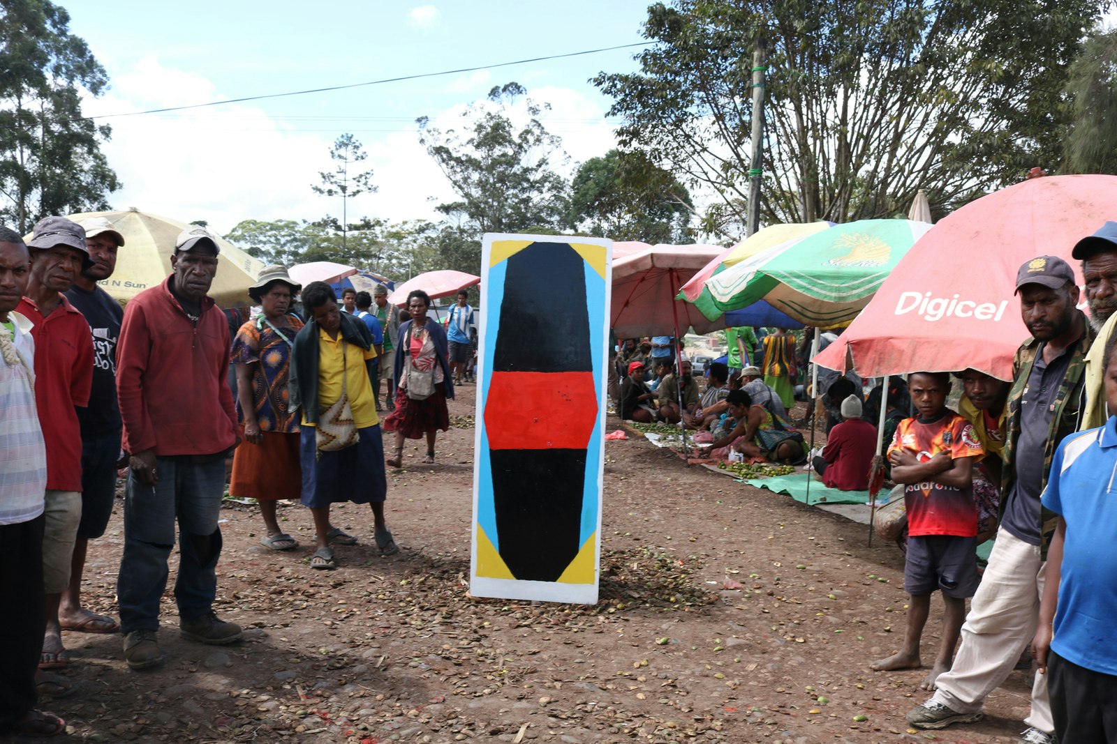 A kuman (painted with a large black shape bisected by a red rectangle), is positioned in the thoroughfare of a market, with patrons standing on each side of it and staring directly at us. Most of them are frowning. 
