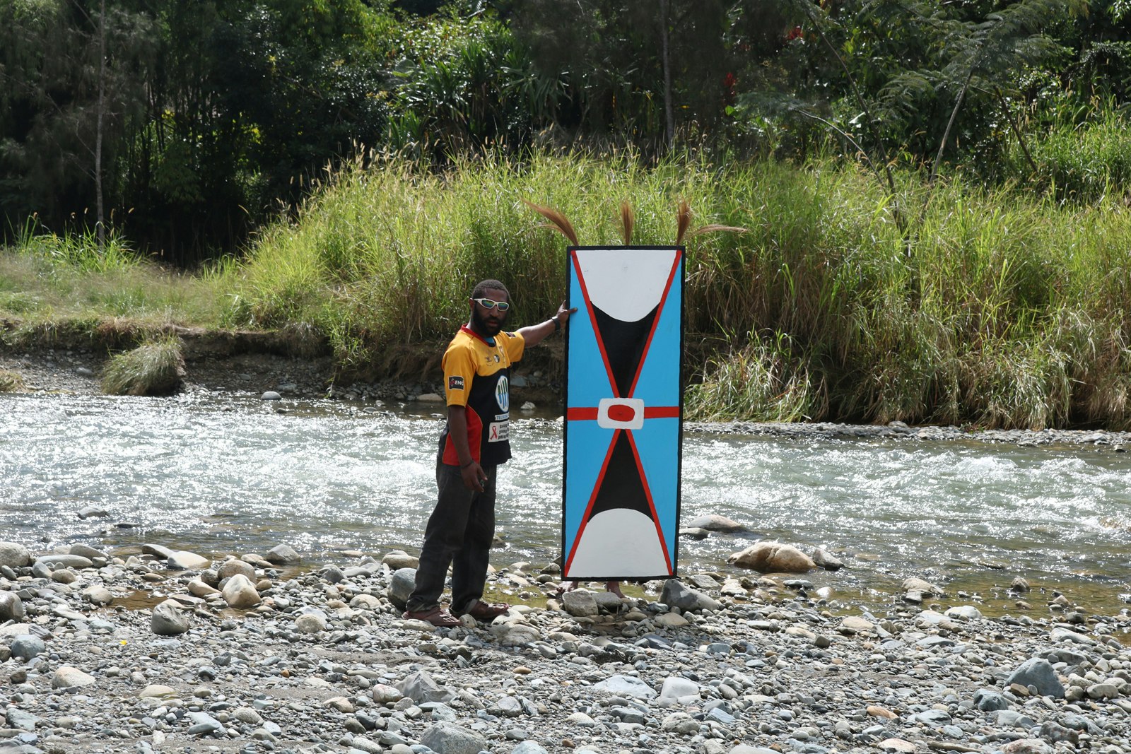 A dark-skinned Pasifika man wearing a jersey, trousers and sunglasses stands on a rocky riverbed, holding up a blue kuman.