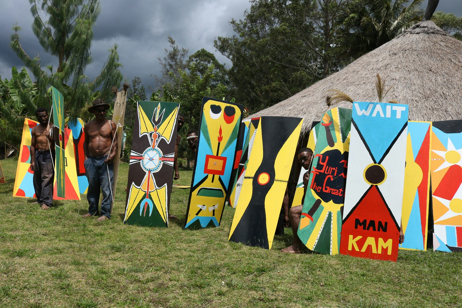 A series of kuman, painted with bright yellow triangles and blue and red shapes, outside a grass hut. One of them is painted with the words, Wait Man Kam.