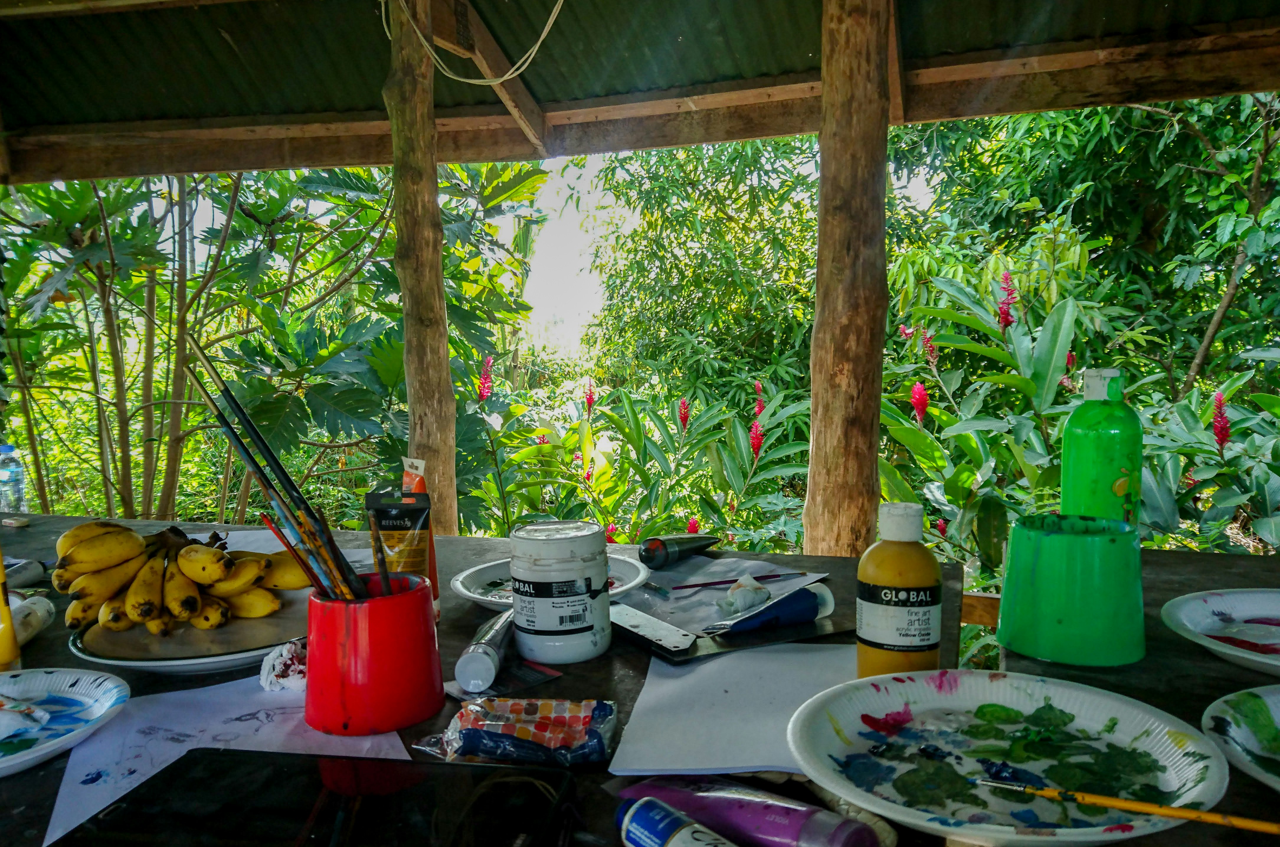 A table laid with paints, palettes and paintbrushes set up outdoors under an awning, surrounded by lush green tropical vegetation.