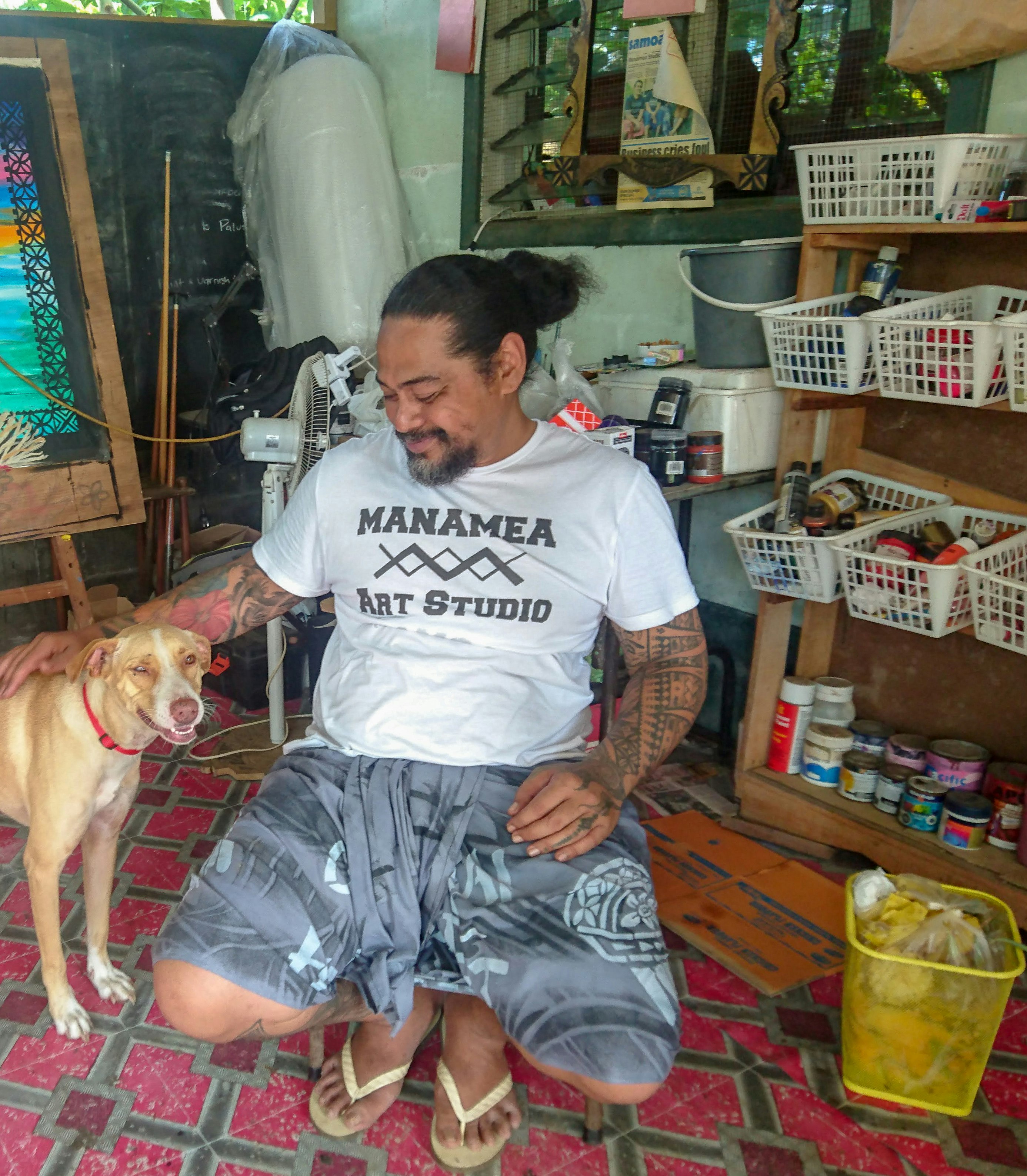 A Pasifika male-presenting figure wearing a white shirt printed with 'Manamea Art Studio', sitting down and patting a dog next to him. On his left are storage containers filled with paints and other art supplies.