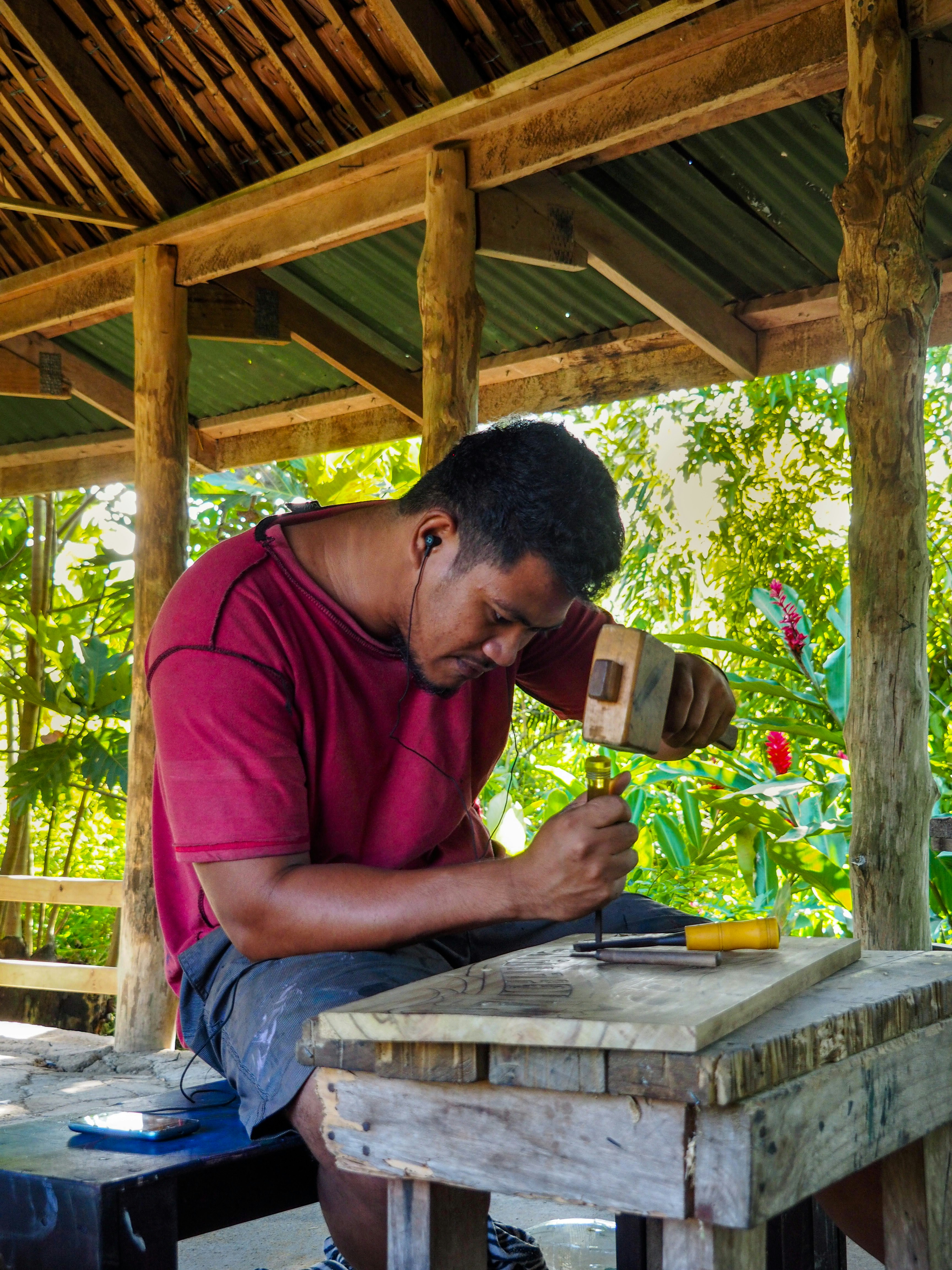 A Samoan male-presenting figure in a maroon t-shirt and navy work shorts leans over a table with a chisel and mallet, wearing earphones as he works.