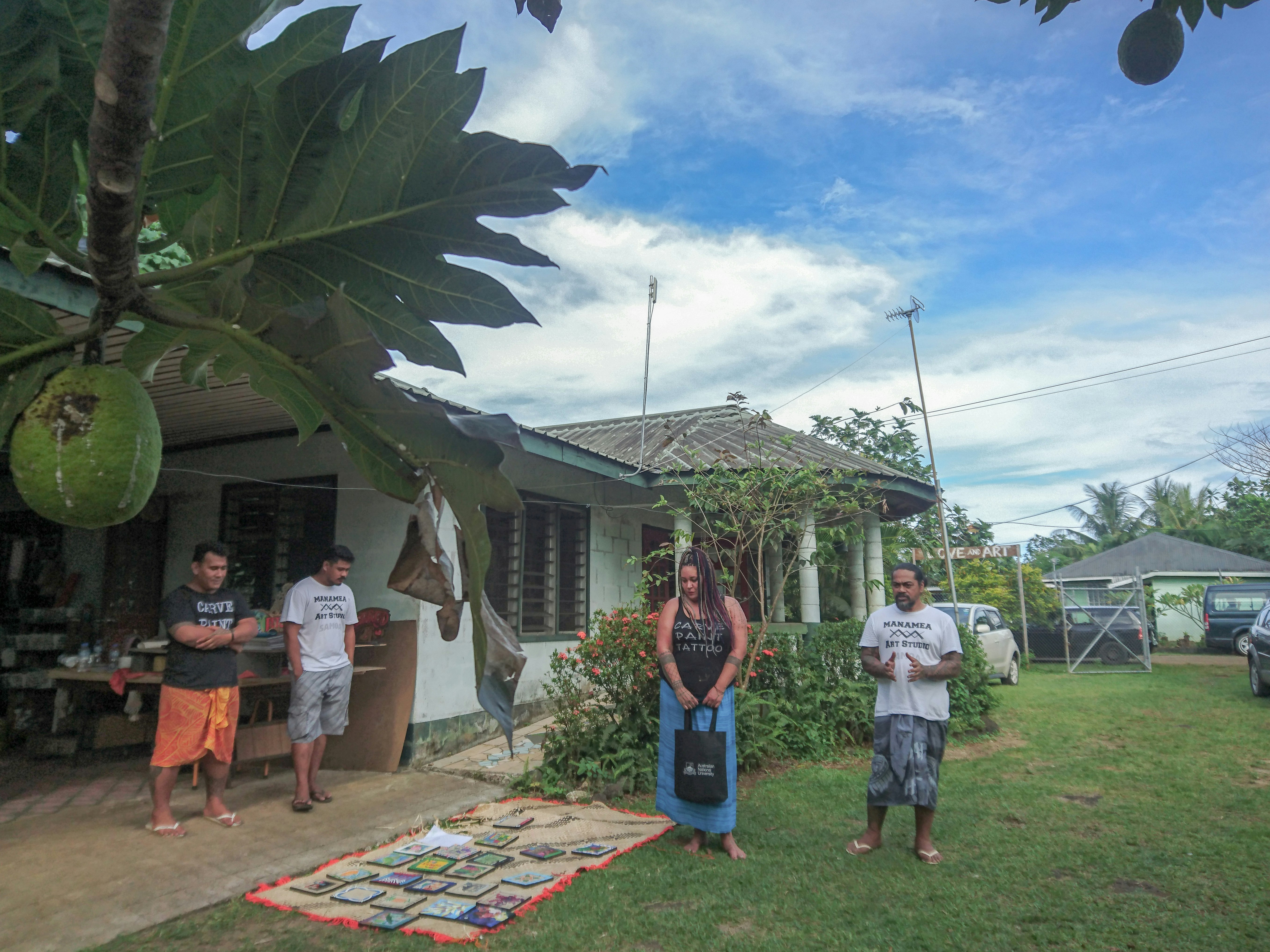 Three male-presenting figures and a female-presenting figure standing on the lawn of a house in Samoa, near a rug laid with framed art.