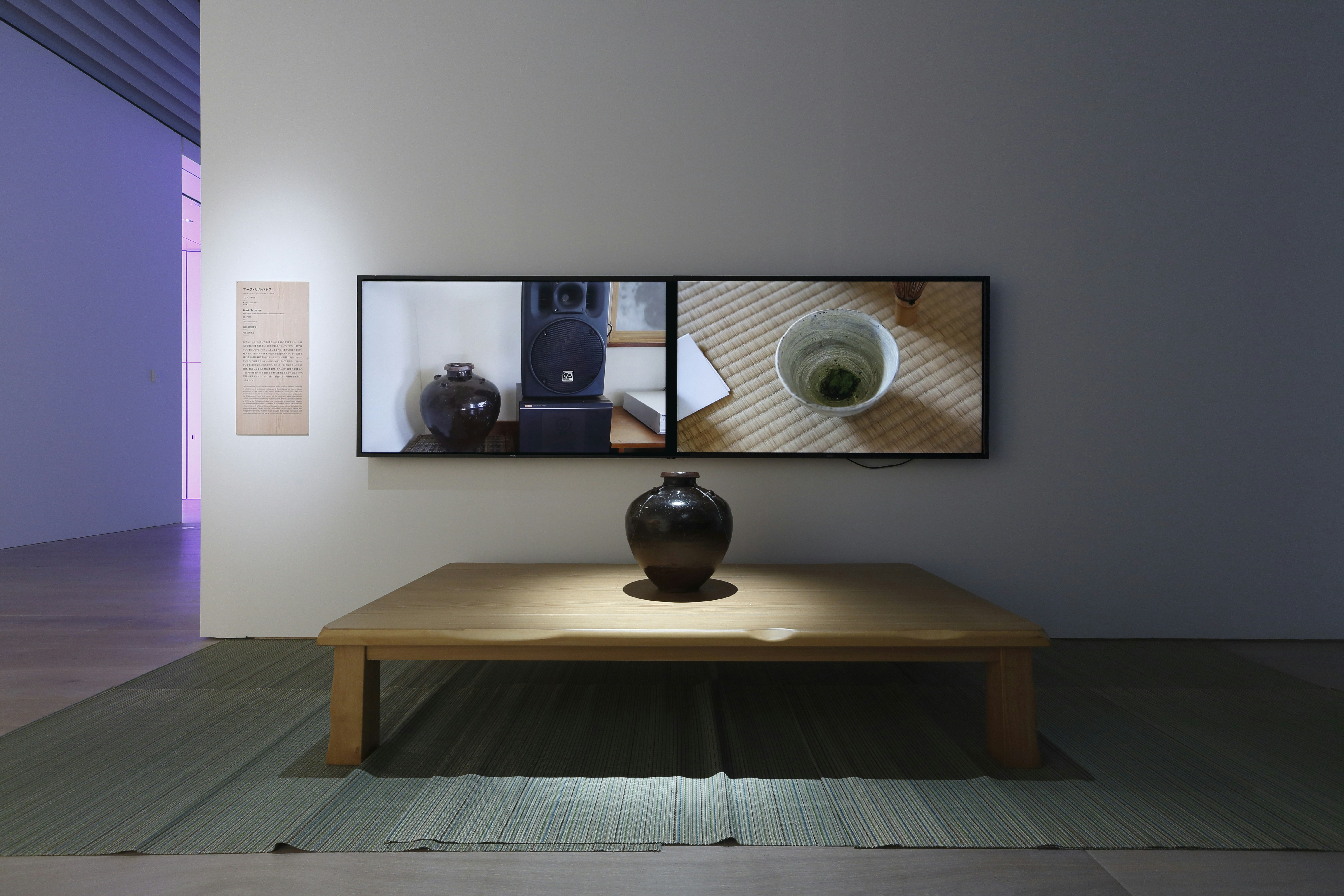A black vase positioned in the middle of a wooden platform with a sole spotlight on it. Behind are two TV monitors, one showing the black vase next to a speaker, the other showing the inside of a teacup on a tatami mat.