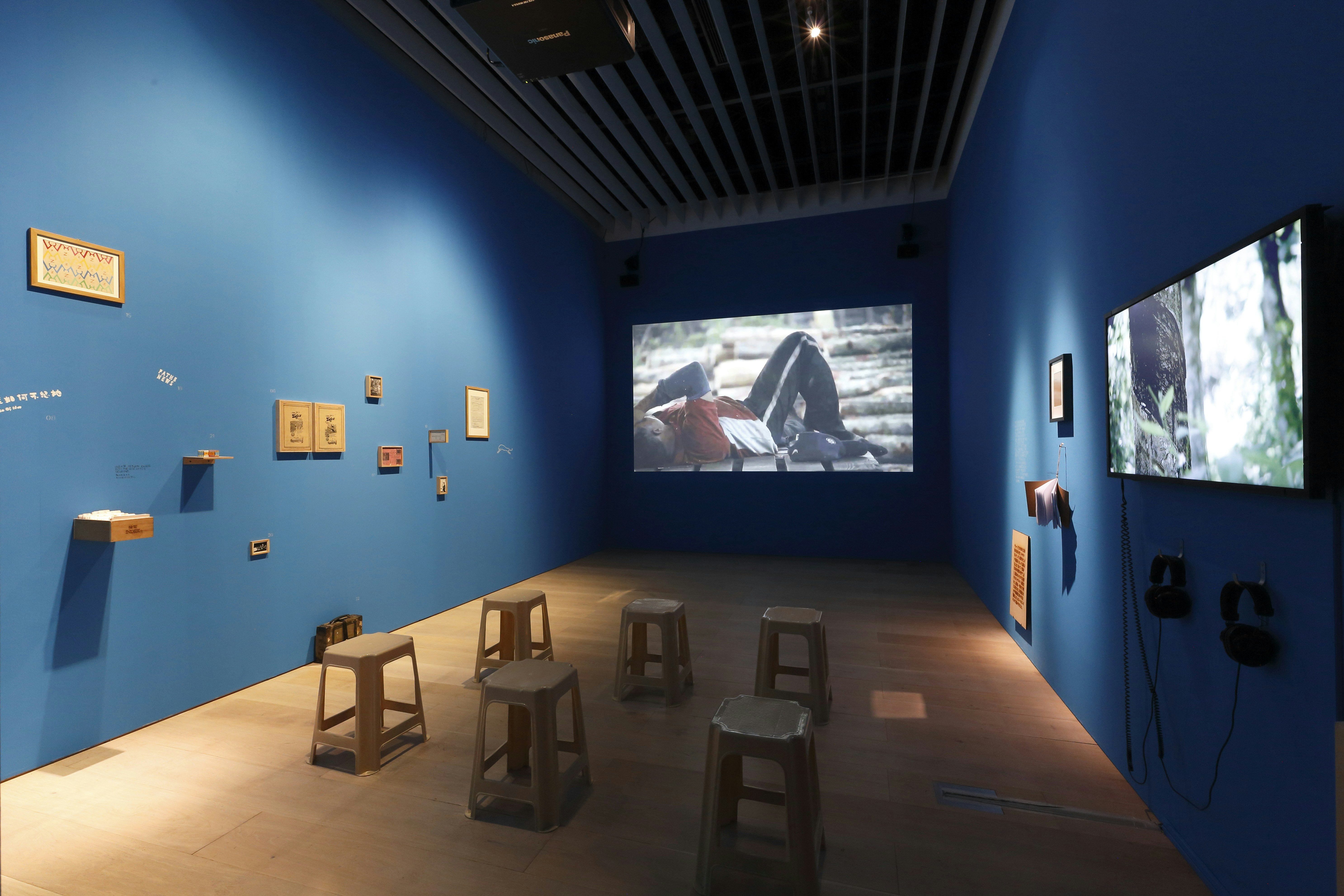 A blue, dimly lit room with wooden pieces installed on the left wall, a projection screening at the back wall, and a TV monitor with attached headphones installed on the right wall. There are six wooden stalls arranged in the middle of the room, facing the projection.
