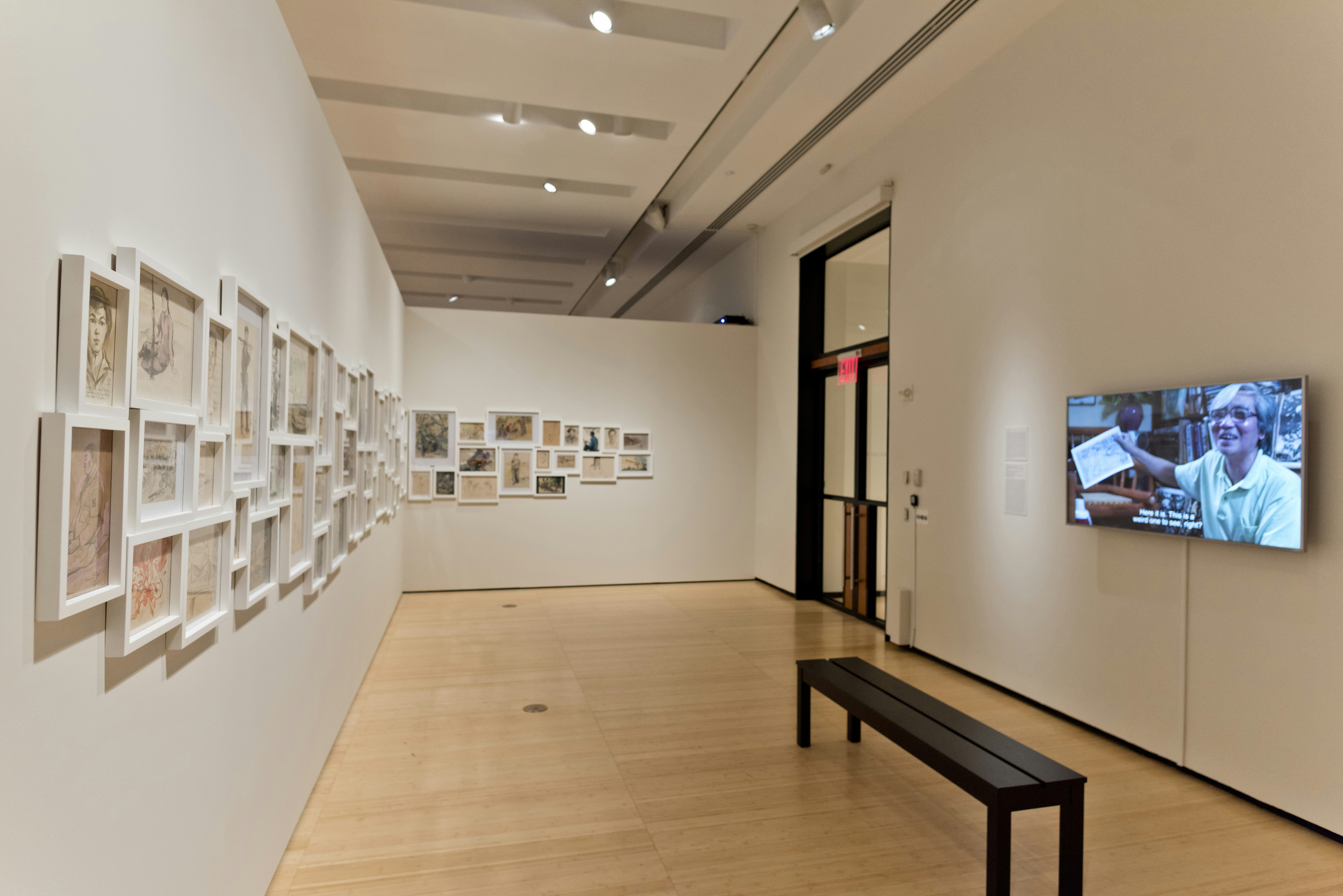 Framed sketches installed across two walls in an art gallery, facing a mounted television monitor showing a Vietnamese male-presenting figure holding up a sketch.