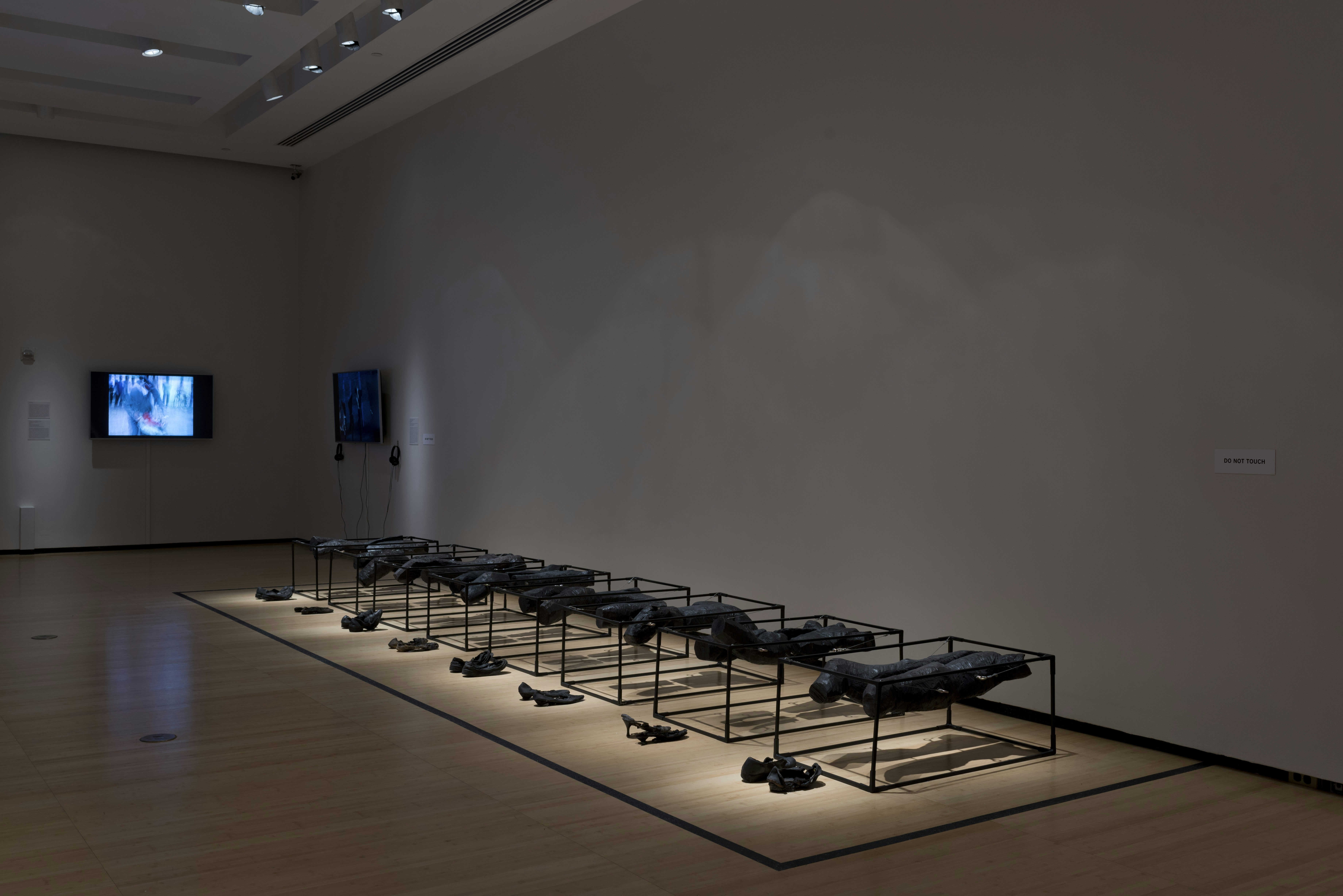 Hunks of burned wood laid on metal frames on a gallery floor, with a pair of black shoes placed in front of each frame. In the corner are two television monitors, one with headphones attached.