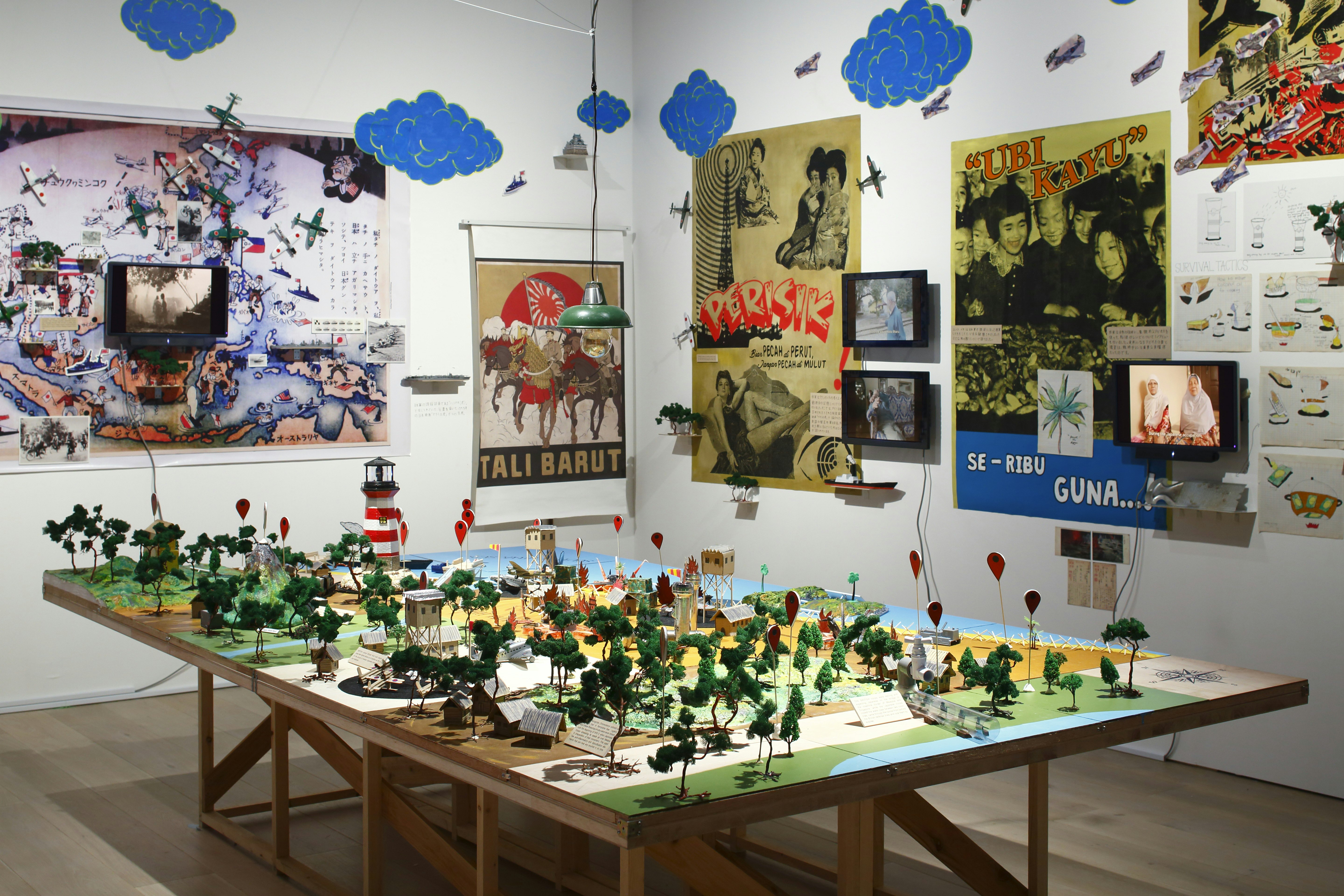 A miniature scene of a forest, drop pins, viewing towers and a lighthouse set up on a drafting table. On the walls are colourful pop-art style posters printed with Indonesian slogans. Blue cloud decals and stickers of military aircraft are stuck on the walls.   