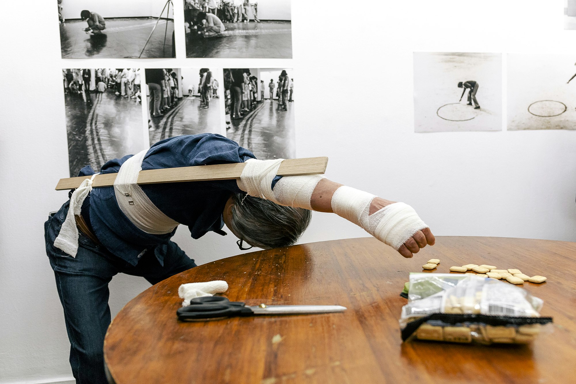 A silver-haired man with a splint bandaged along his torso and up his right arm leans over a table trying to grab some small biscuits laid out.