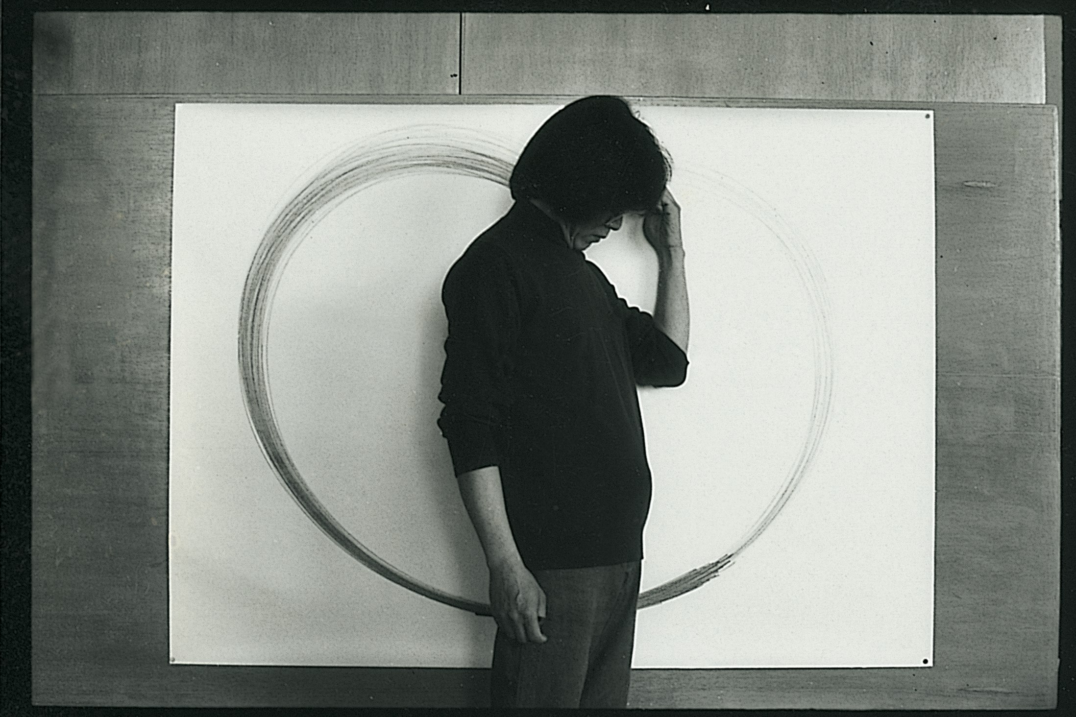 Side profile of a young Lee Kun-Yong, with shoulder length black hair and a black sweater, stands against a white canvas with a circular brushstroke. He is looking downwards with his hand lifted to his forehead.