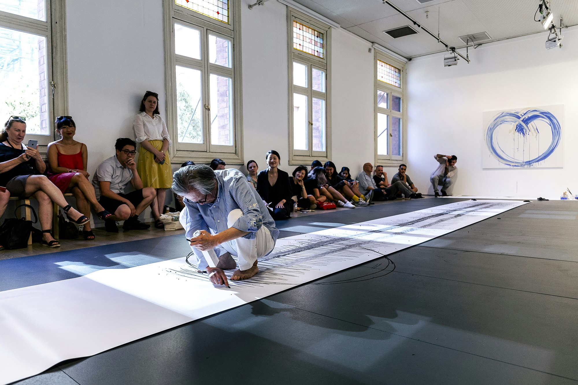 Lee Kun Yong, a Korean male-presenting figure with silver hair, a blue striped shirt and white paints squats barefoot on a long scroll of white paper, drawing with a stick of charcoal.