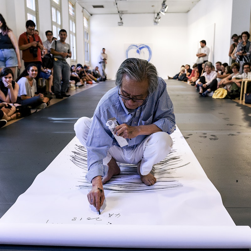 Lee Kun-Yong, an East Asian male-presenting figure in a striped blue shirt and white pants squats on a long scroll of white paper, scribbling horizontally with a stick of charcoal. On either side of him is a crowd of onlookers, some of whom are holding up phone cameras.