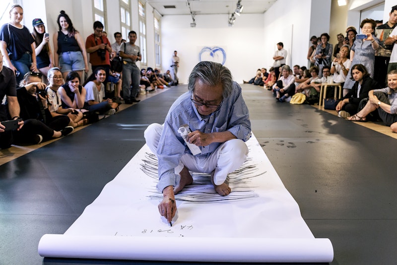 Lee Kun-Yong, an East Asian male-presenting figure in a striped blue shirt and white pants squats on a long scroll of white paper, scribbling horizontally with a stick of charcoal. On either side of him is a crowd of onlookers, some of whom are holding up phone cameras.