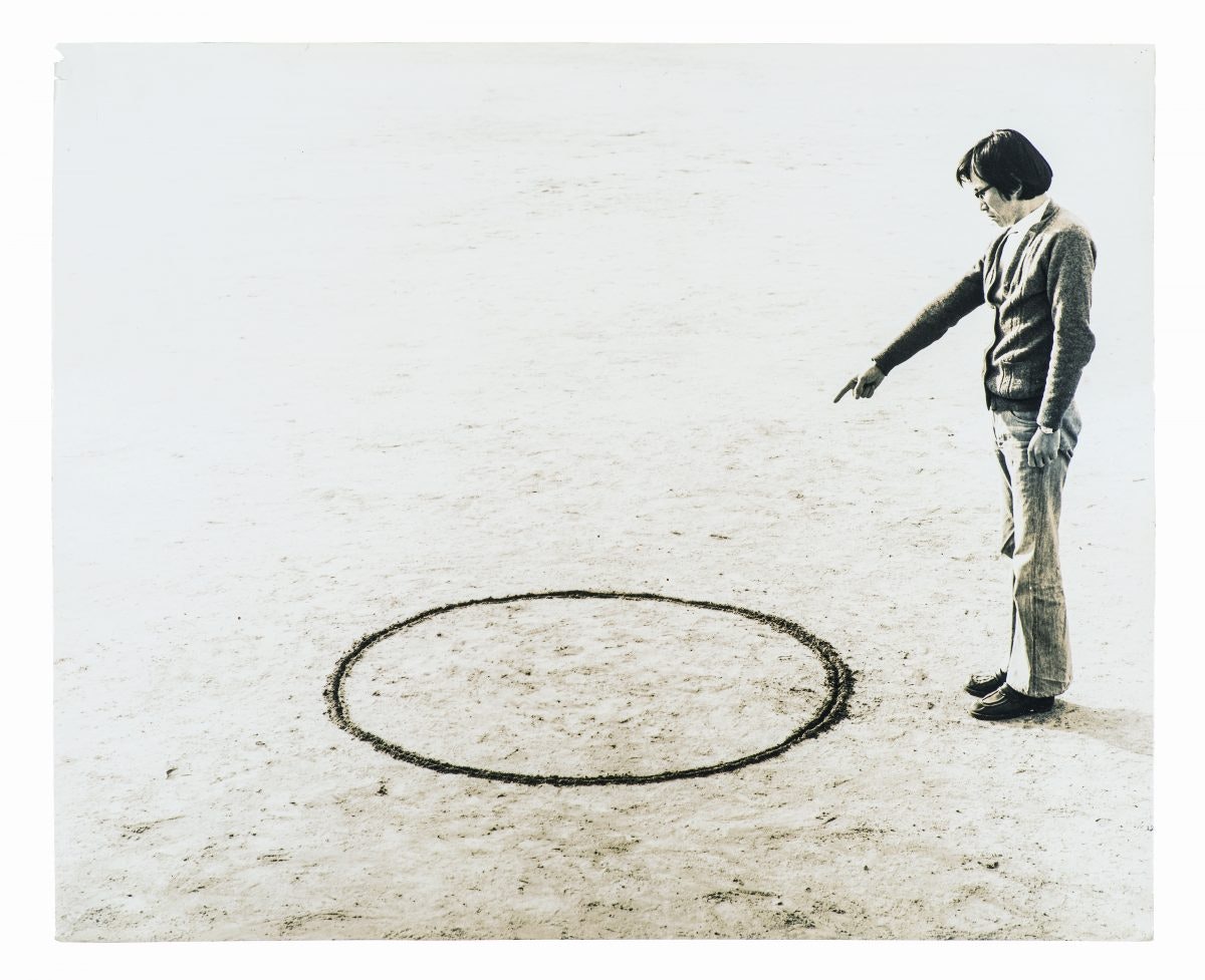 A Korean male-presenting figure with a dark black bob, wearing glasses, a button-up cardigan and slightly flared trousers, stands while pointing to a circle drawn in the ground.