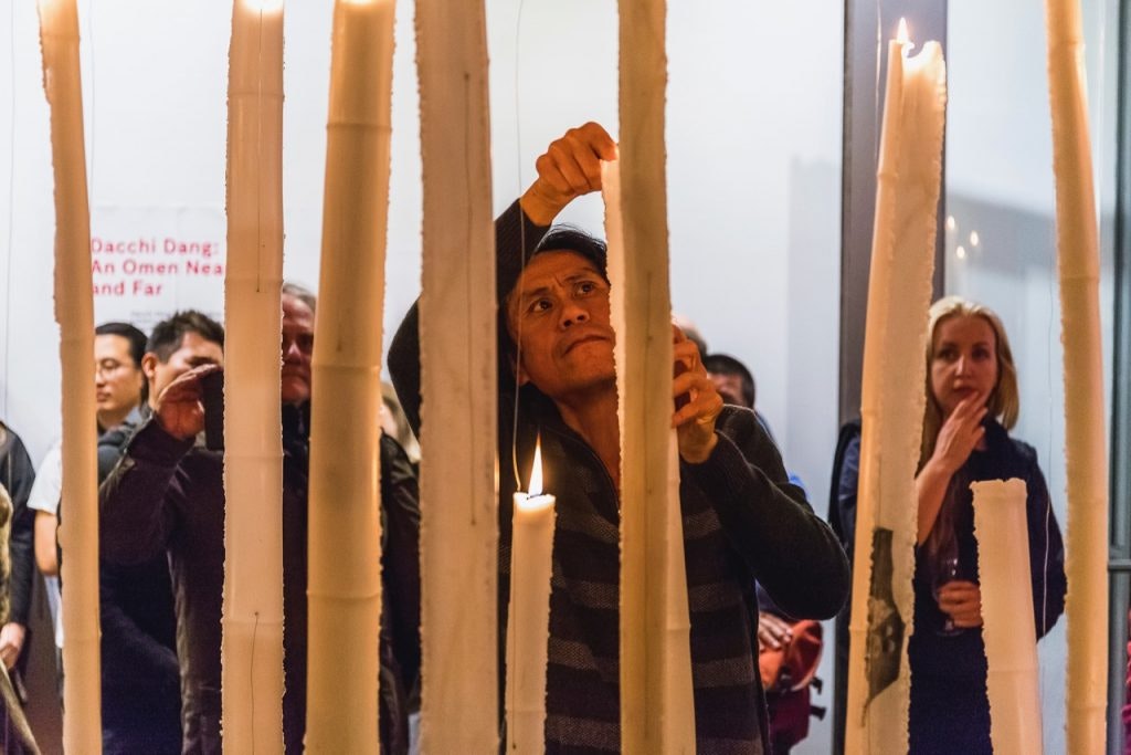 Dacchi Dang, a tanned Southeast Asian man, lights a series of tall candles in front of an audience at 4A.