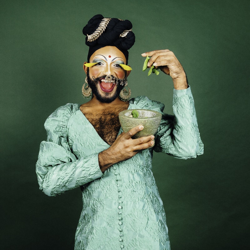 Radha, a South Asian drag performer, smiles at the camera while wearing a turquoise satin dress and black turban, with silver eyeshadow, thinly drawn brows and extravagant yellow lashes. They are holding a mortar bowl in one hand and a few mint leaves in the other.