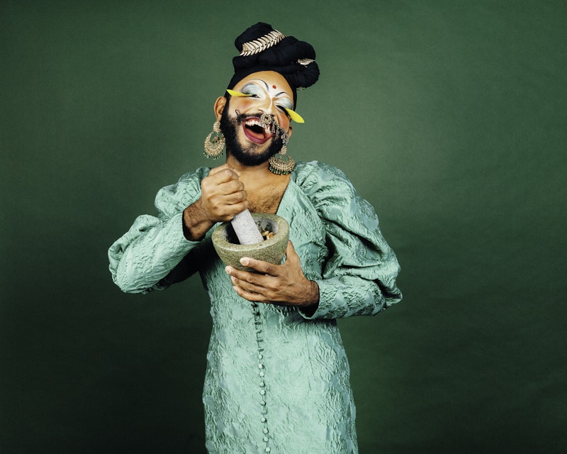 Radha, a South Asian drag performer, laughs at the camera while grinding ingredient in a mortar and pestle. They wear a turquoise satin dress, gold earrings, yellow false lashes, silver eyeshadow and a black turban.