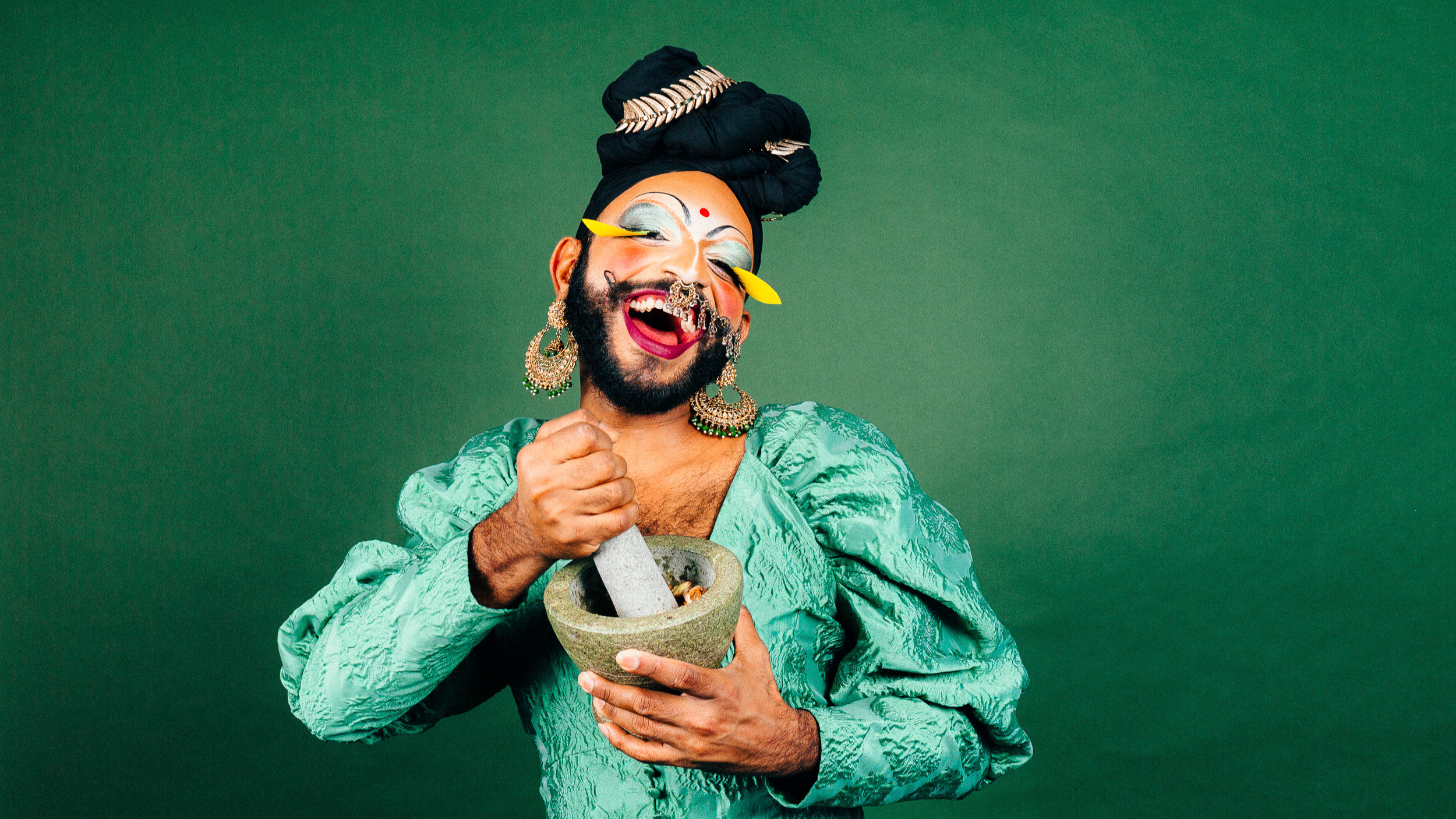 Radha, a South Asian drag performer, laughs at the camera while grinding ingredients in a mortar and pestle. They wear a turquoise satin dress, gold earrings, yellow false lashes, silver eyeshadow and a black turban.