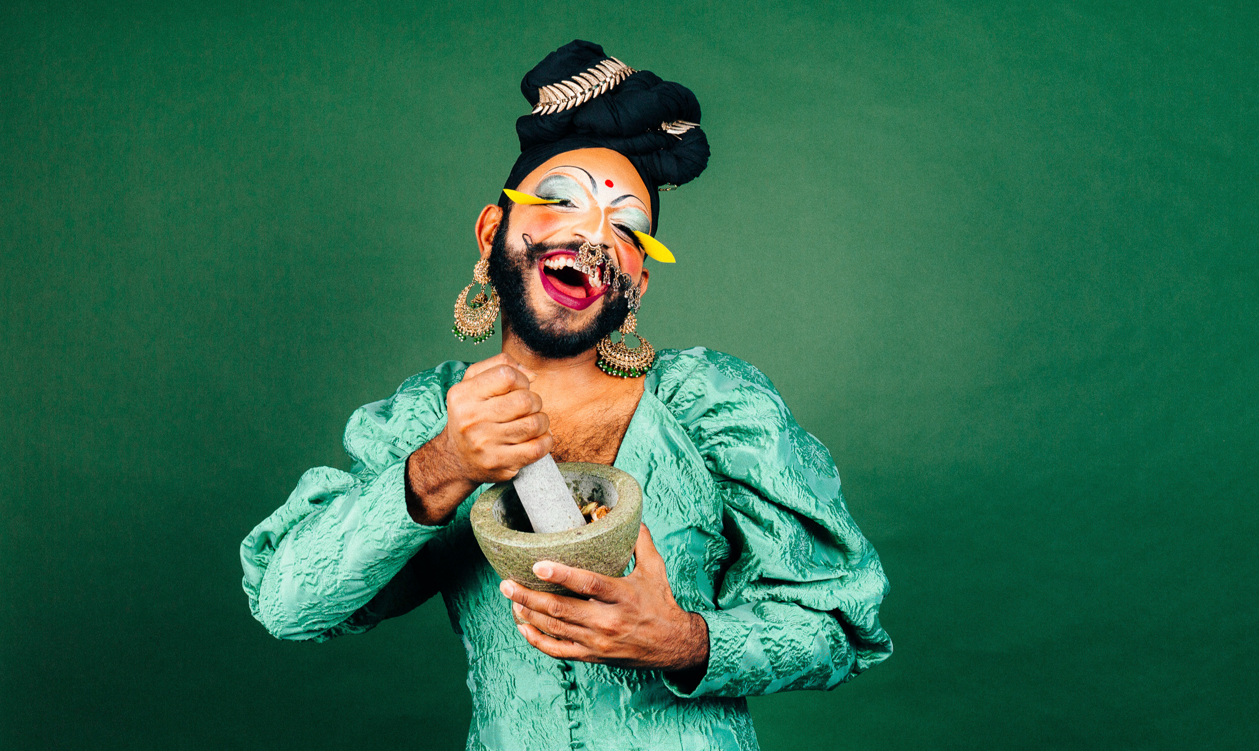 Radha, a South Asian drag performer, laughs at the camera while grinding ingredients in a mortar and pestle. They wear a turquoise satin dress, gold earrings, yellow false lashes, silver eyeshadow and a black turban.