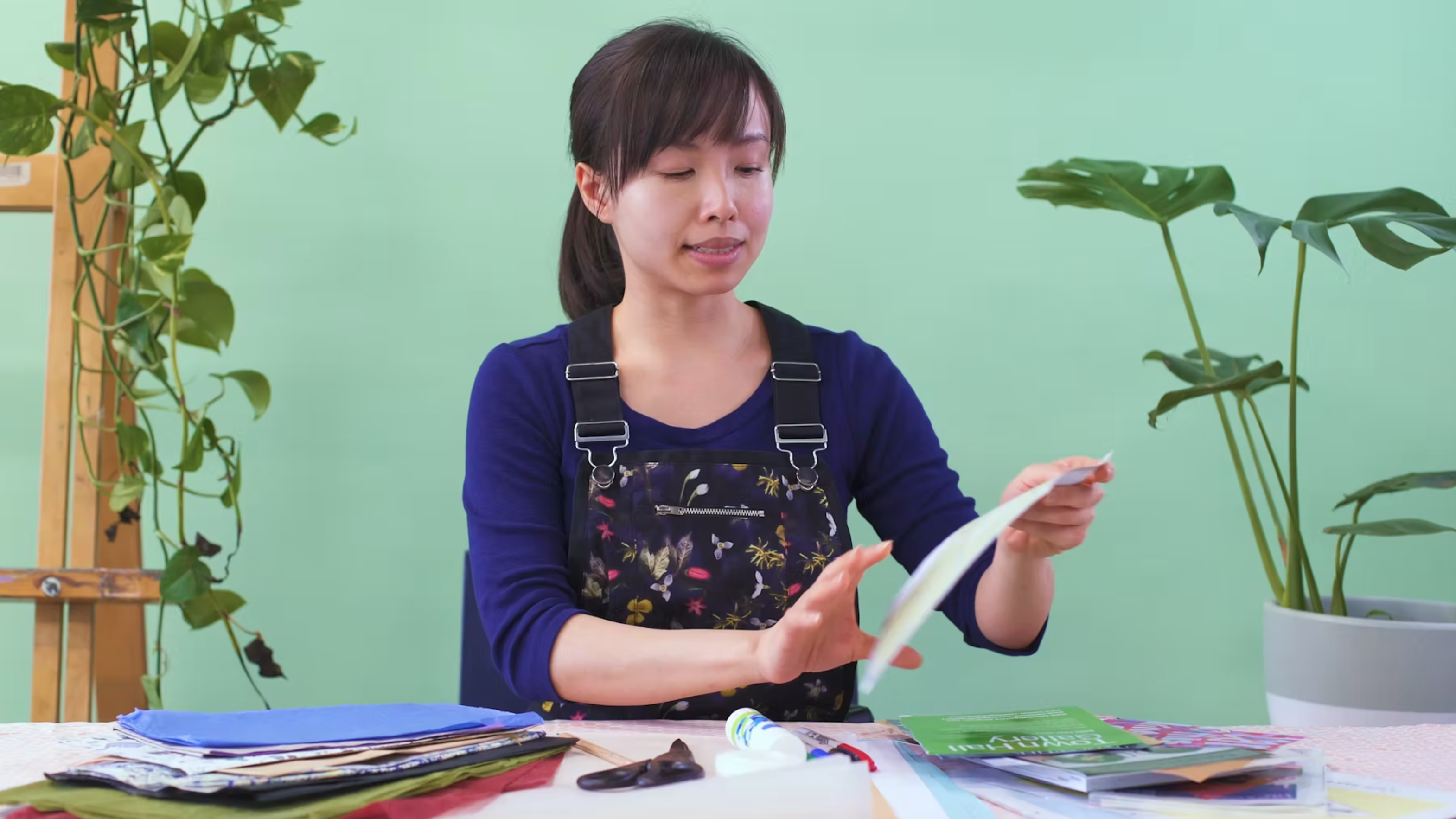 An East Asian female-presenting artist wearing overalls cuts paper in a room with teal walls and vibrant indoor plants.
