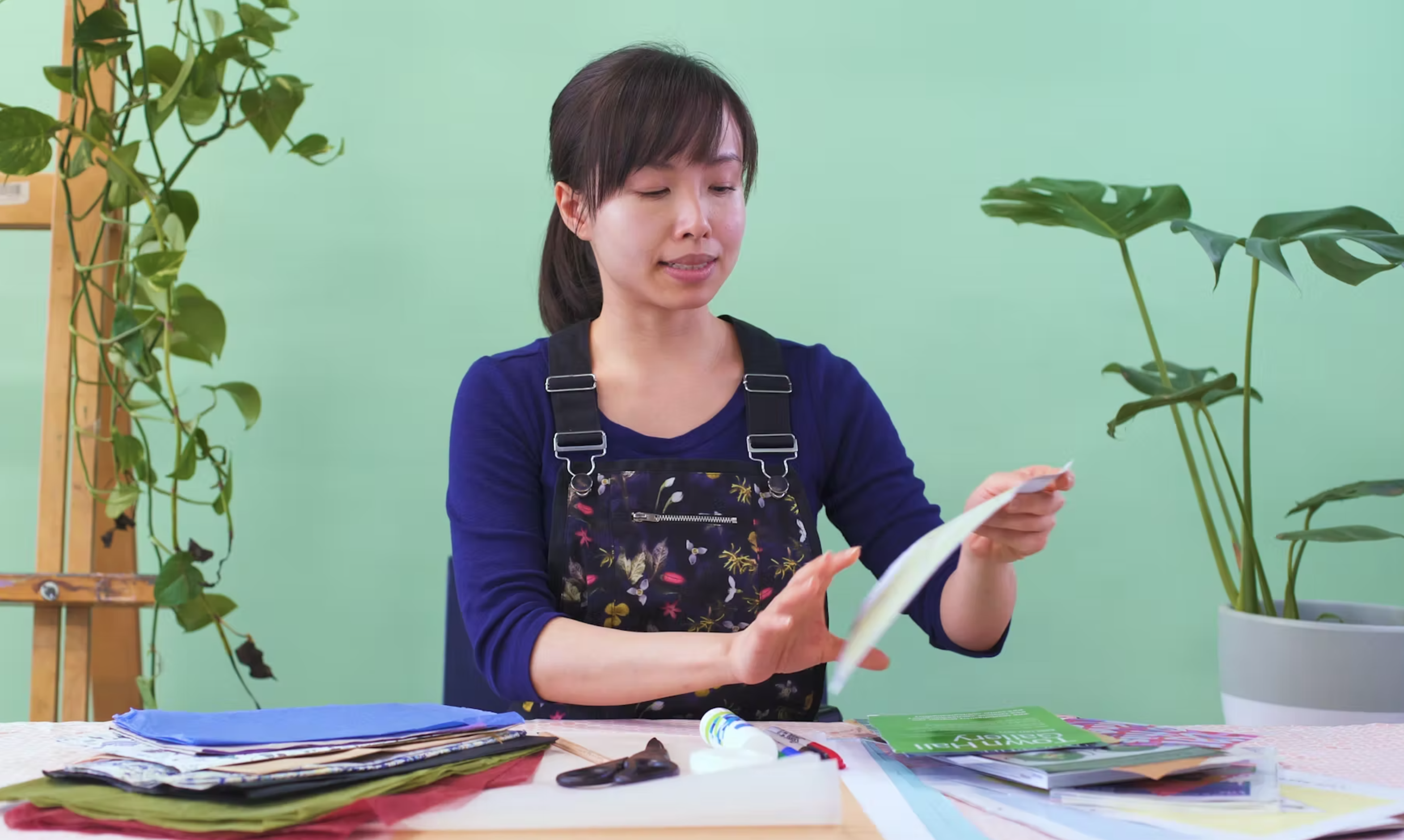 An East Asian female-presenting artist wearing overalls cuts paper in a room with teal walls and vibrant indoor plants.