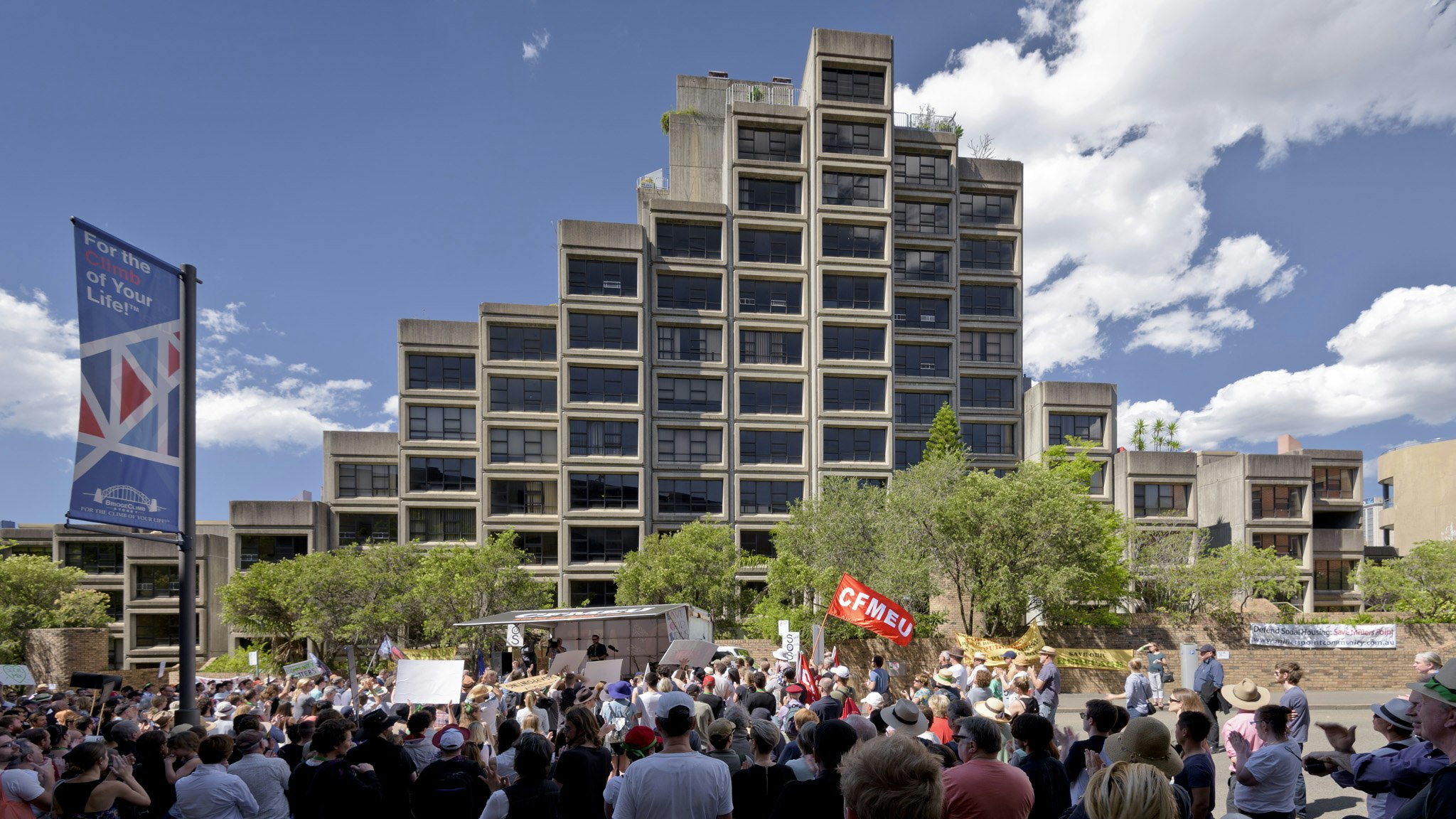 A crowd rallying in front of a housing estate. Someone holds up a flag with the initialism, CFMEU.