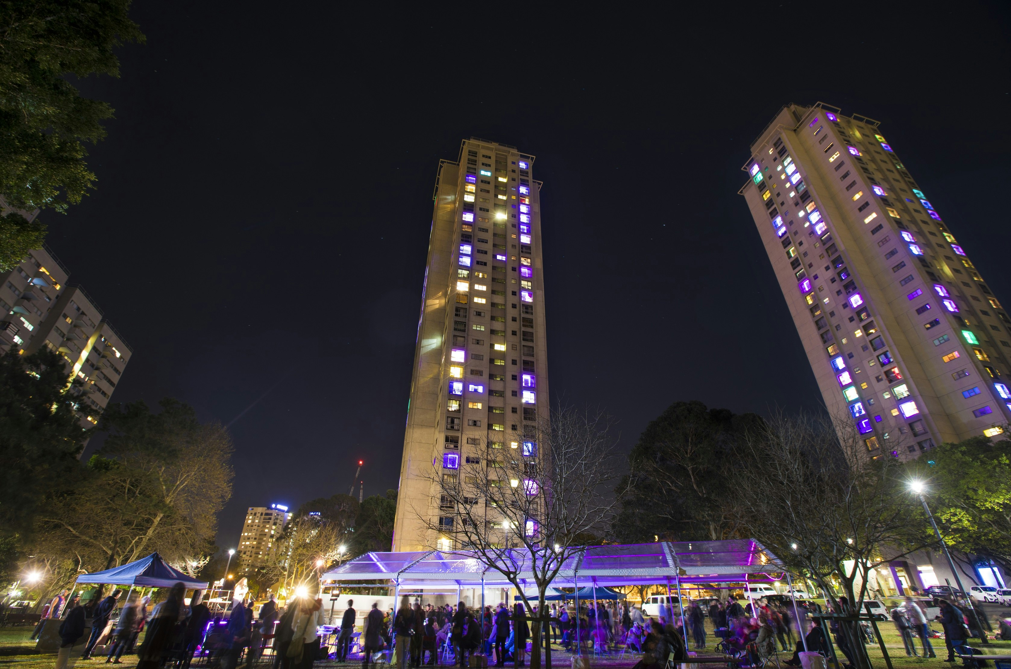 Two apartment towers with neon purple, green and orange windows lit up at night.